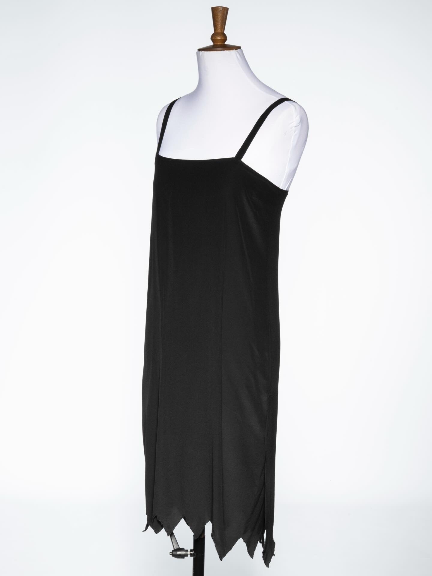 A black sleeveless dress displayed on a mannequin. The dress features a straight cut with a hemline that has a jagged pattern, giving it a distinct appearance.
