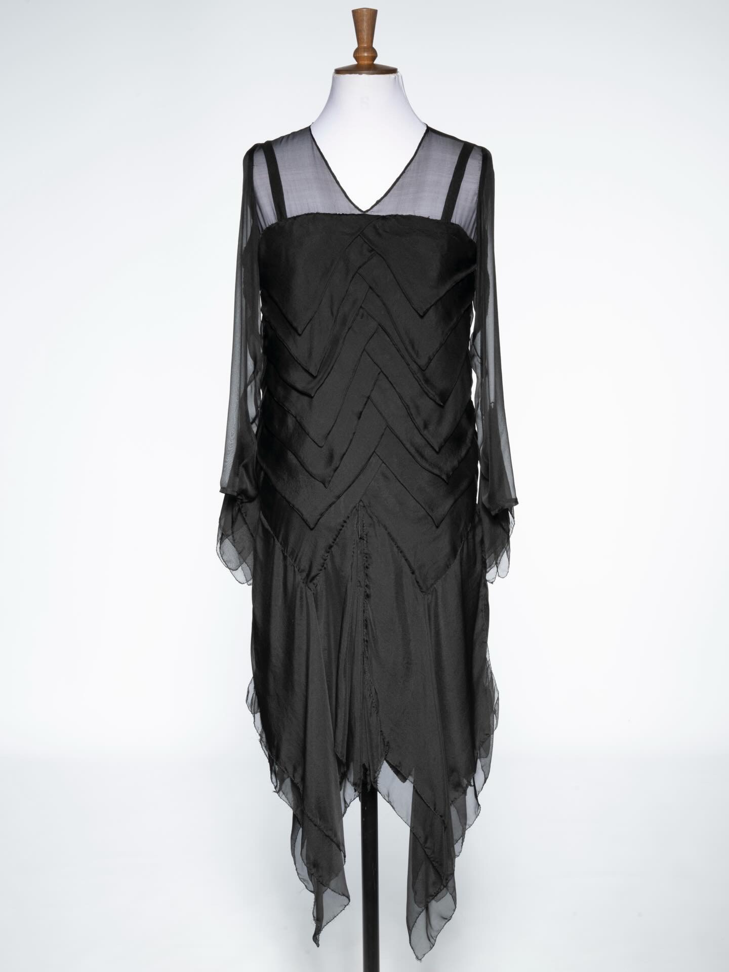 A black dress with a V-neckline and long, sheer sleeves displayed on a mannequin. The dress features a zigzag pattern and a flowing hemline that is longer at the back, creating an elegant silhouette.