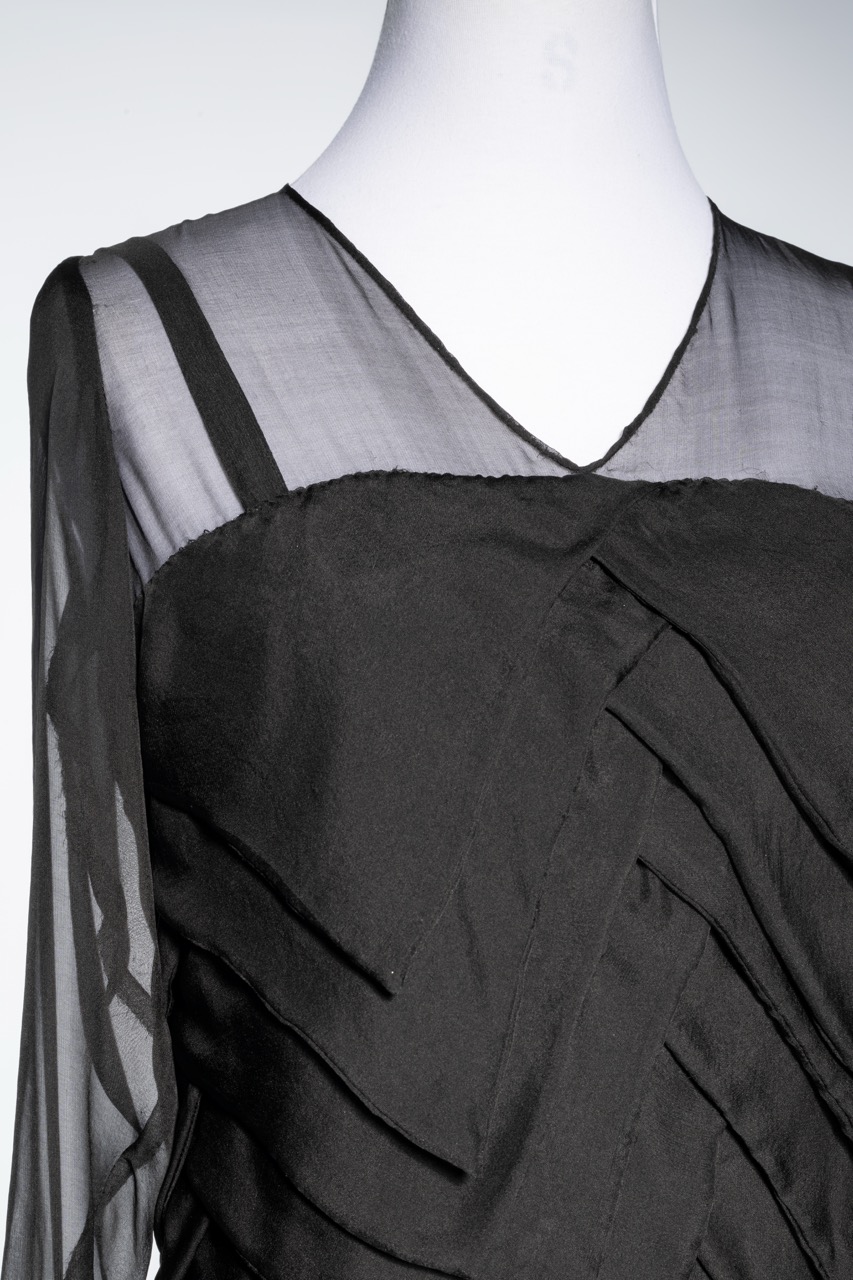 A black dress featuring a layered design with sheer long sleeves and a deep V-neck. The fabric appears smooth and drapes elegantly over the mannequin, emphasising its intricate detailing.