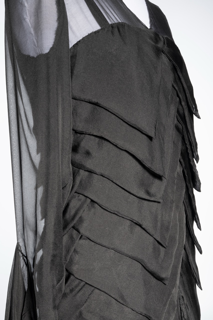 A close-up of a black dress featuring layered ruffle details on its side. The fabric appears silky and smooth, with a sheer overlay visible at the top, showcasing intricate texture and design.
