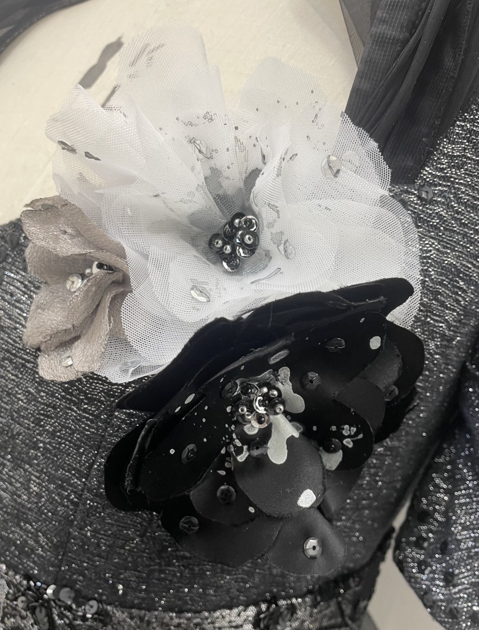 A close-up of a dress embellished with three flowers. The largest flower is black with a glossy finish, and the other two are made of white tulle and a beige fabric, adorned with sequins and beads, all situated on the left side of the neckline.