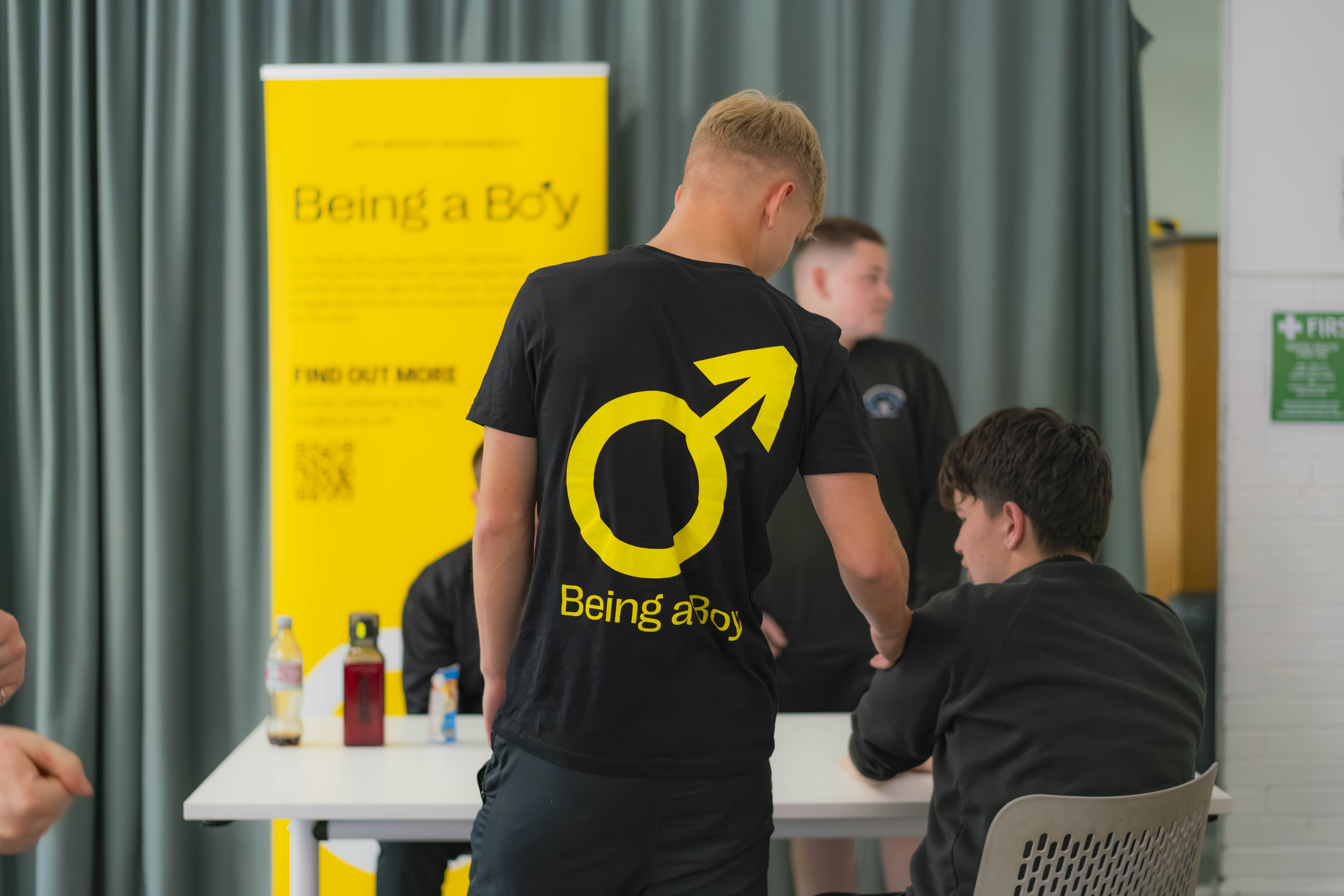 A young person standing and wearing a black T-shirt with a yellow male symbol and "Being a Boy" along the bottom talks with a second young person seated at a table. A yellow banner with "Being a Boy" along the top is visible in the background.