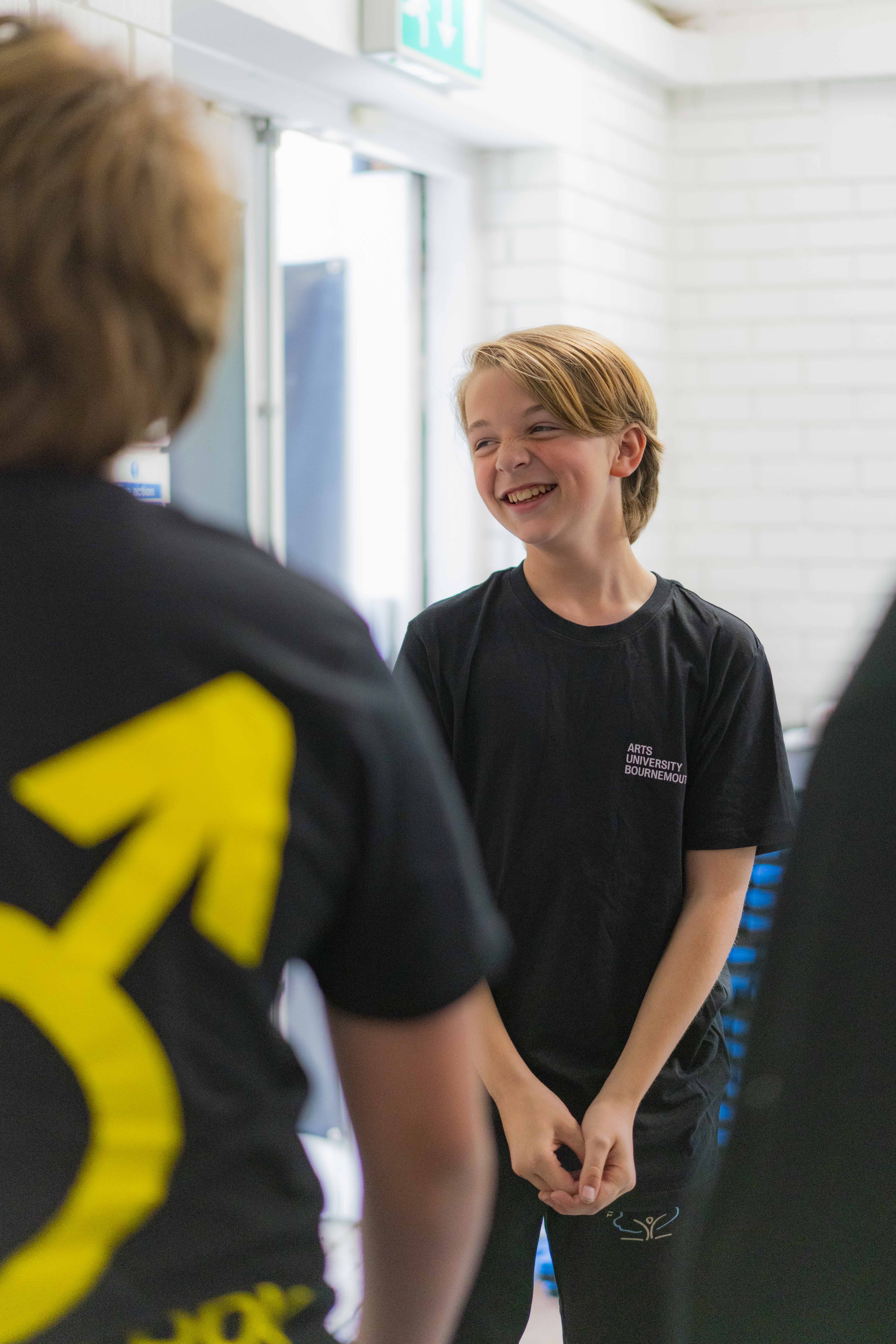 Booy smiling in a white room, person with black t-shirt with yellow male gender icon blurred in foreground.