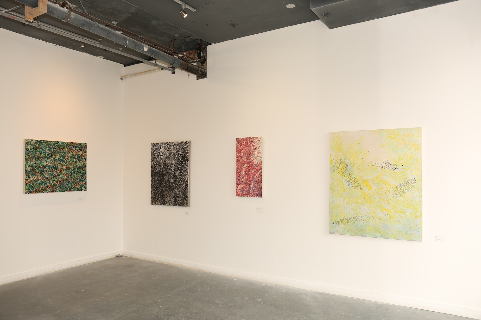Gallery corner with four abstract paintings on white walls: green textured work, dark piece, red composition, and larger yellow work. Exposed ceiling, concrete floor.
