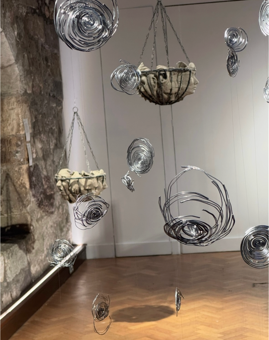 Gallery wall displaying multiple wire basket sculptures and spiral forms in metallic finishes, suspended and mounted against white walls.