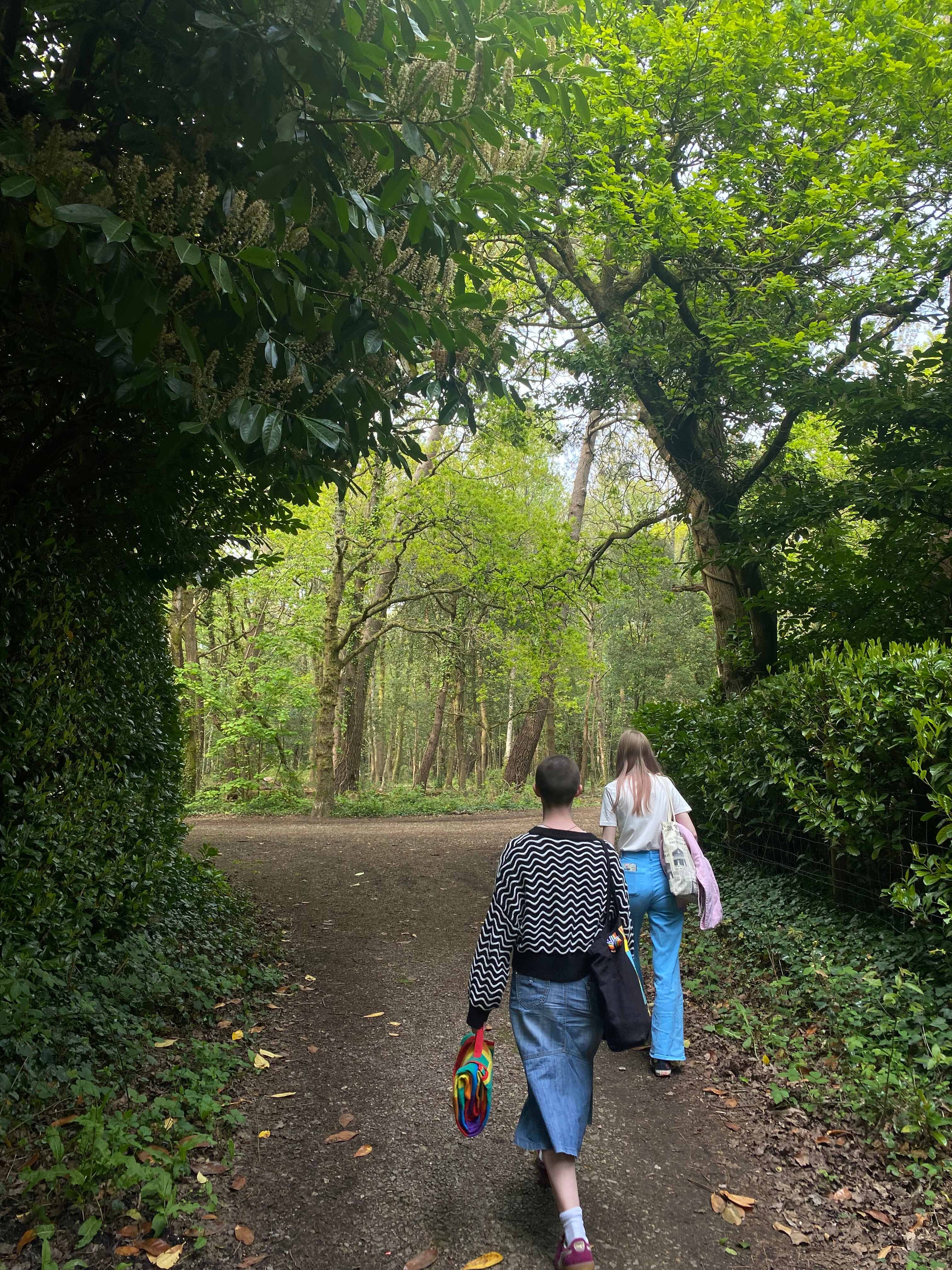 Two people walking on dirt path through green woodland, viewed from behind. Large trees create leafy canopy overhead.
