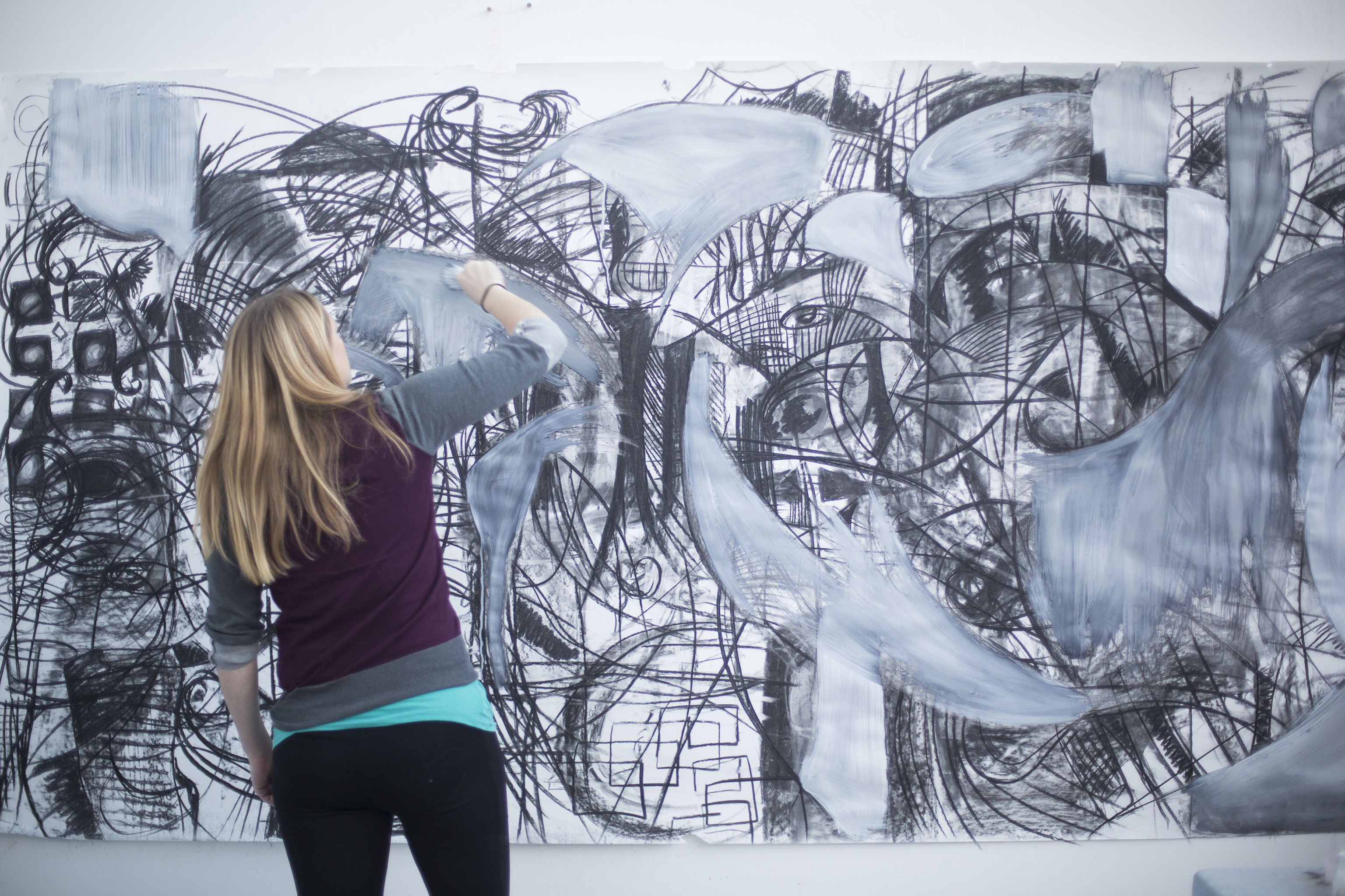 A woman with her back to the camera paints a thick grey brushstroke as part of a black and grey mural.