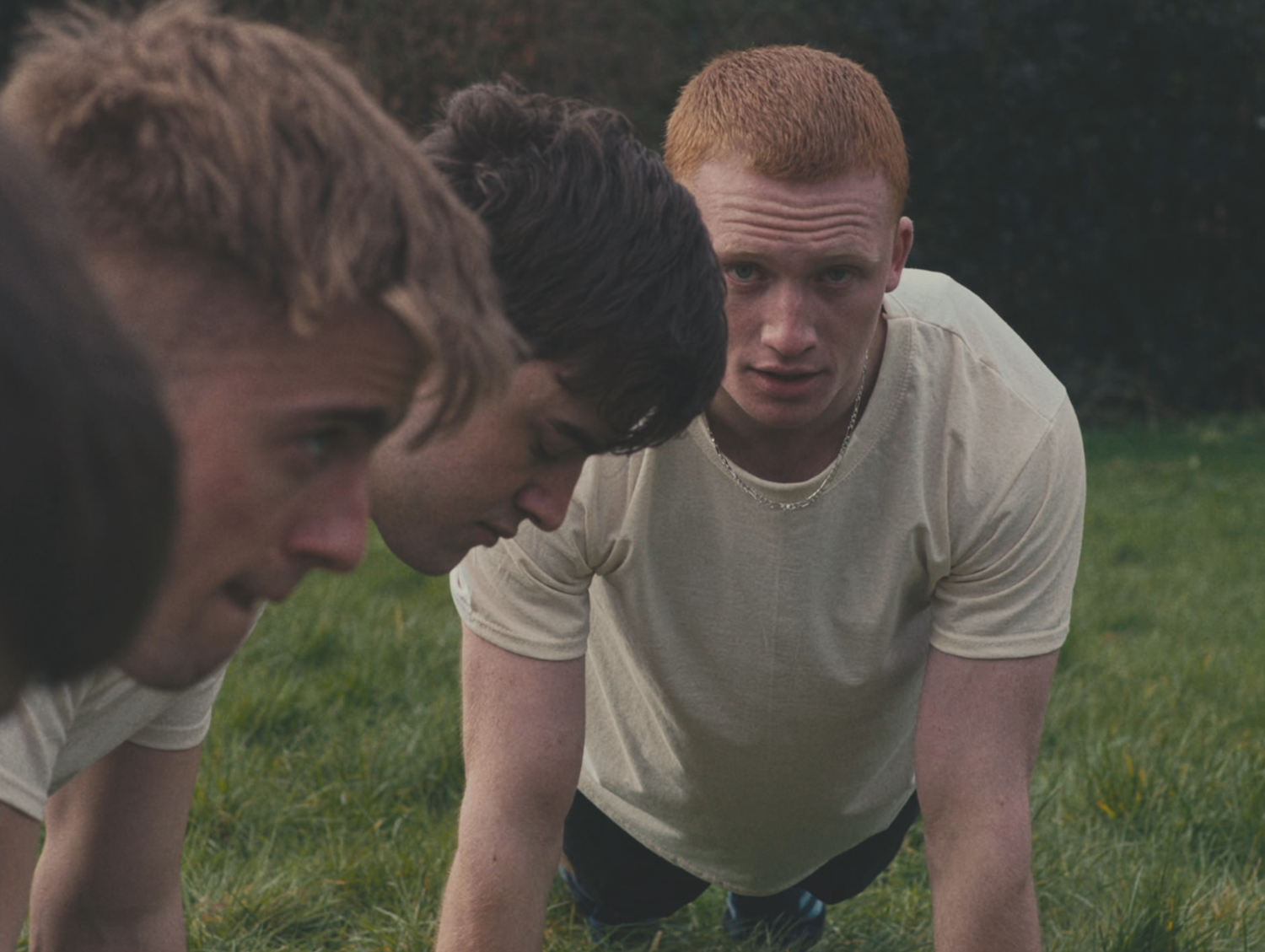 Still shot from Eyes, Front showing characters doing exercise in white t-shirts in a field, one staring directly at the camera.