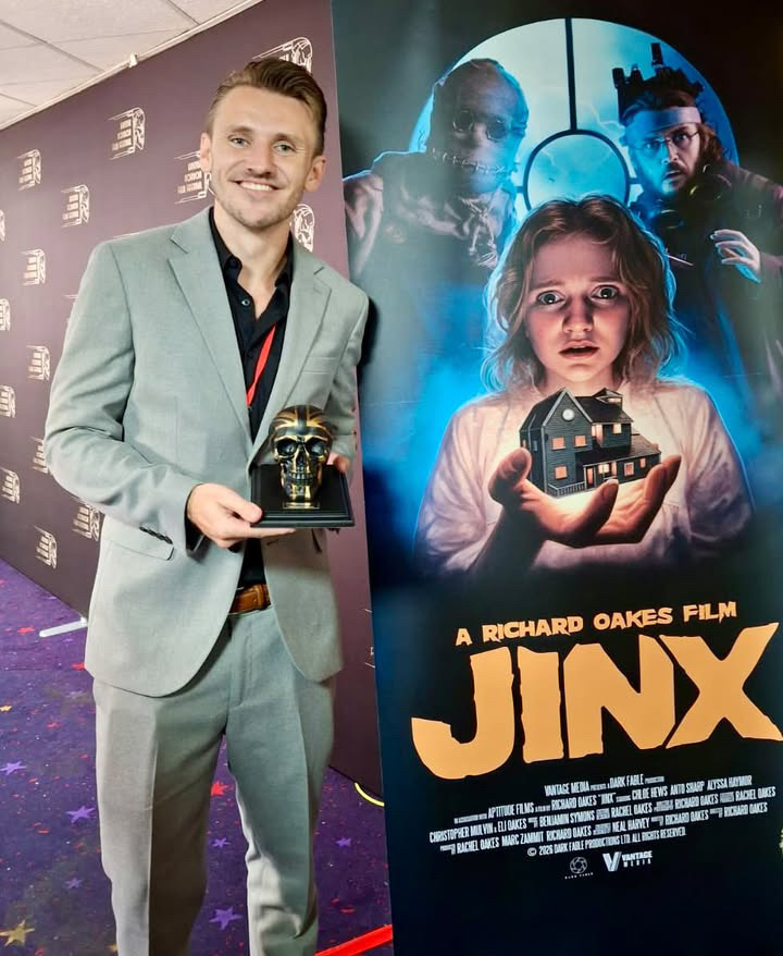 Actor C Mulvin holds a skull-shaped award beside a JINX film poster, with a young girl, two figures and a house illustrated on the banner.