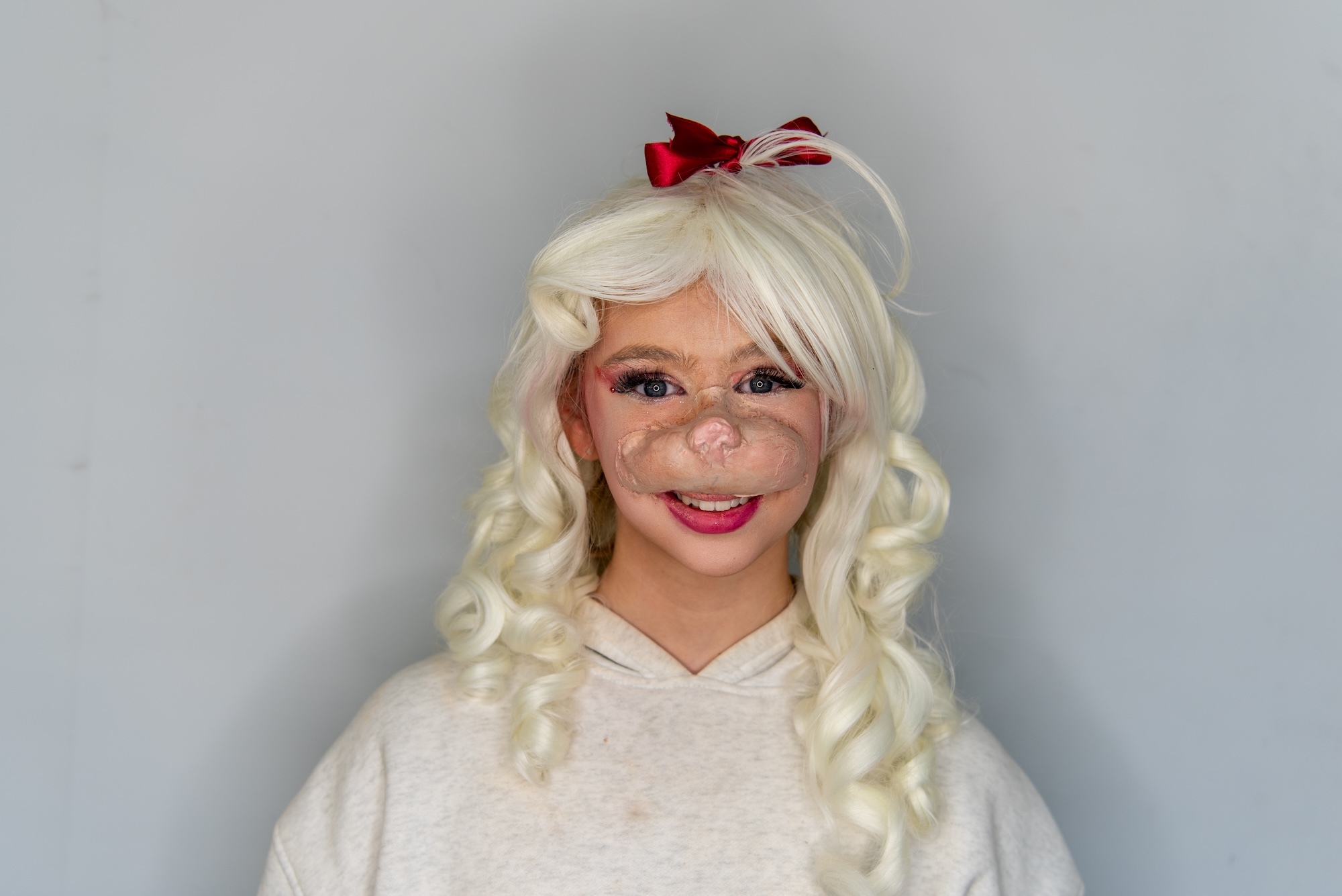 Person wearing blonde curly wig with red bow, white hoodie, and theatrical makeup with exaggerated features against grey background