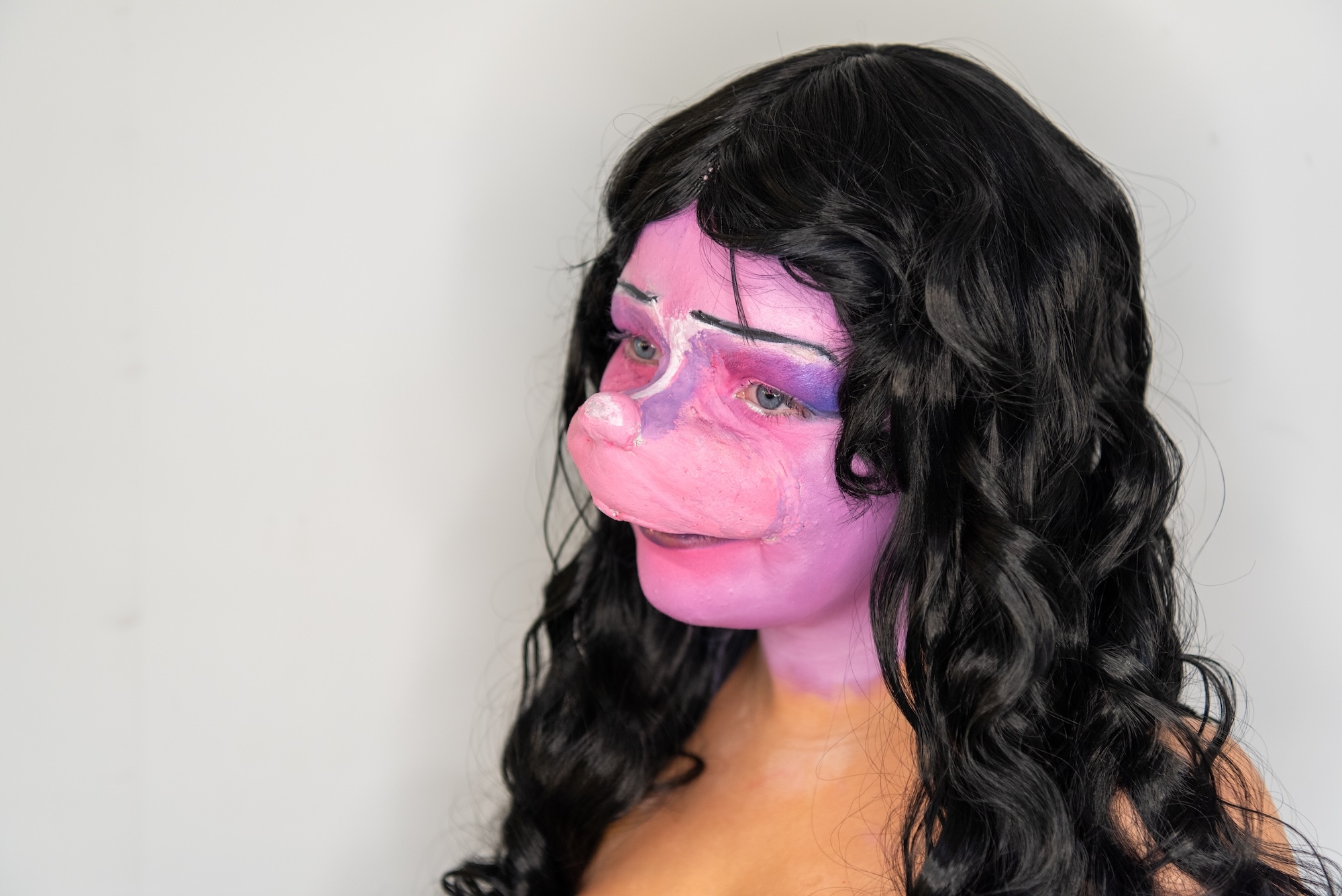 Person with long black curly hair and pink and purple face paint covering left side of face against white background