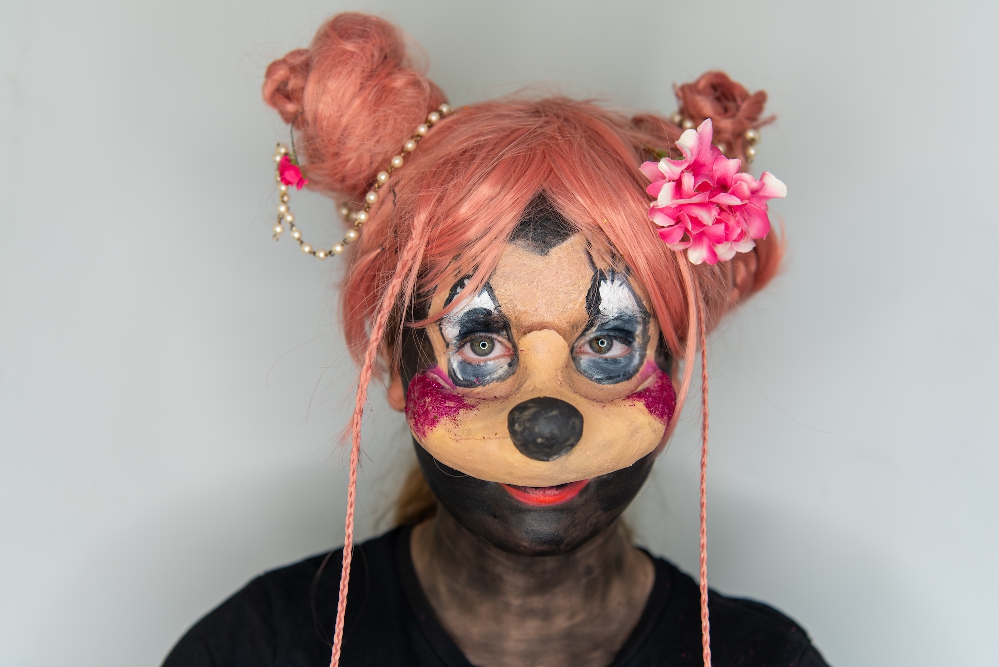 Person wearing dog-like mask with orange/brown face, black nose, blue eyes, reddish hair in buns with pink flowers and beaded chains