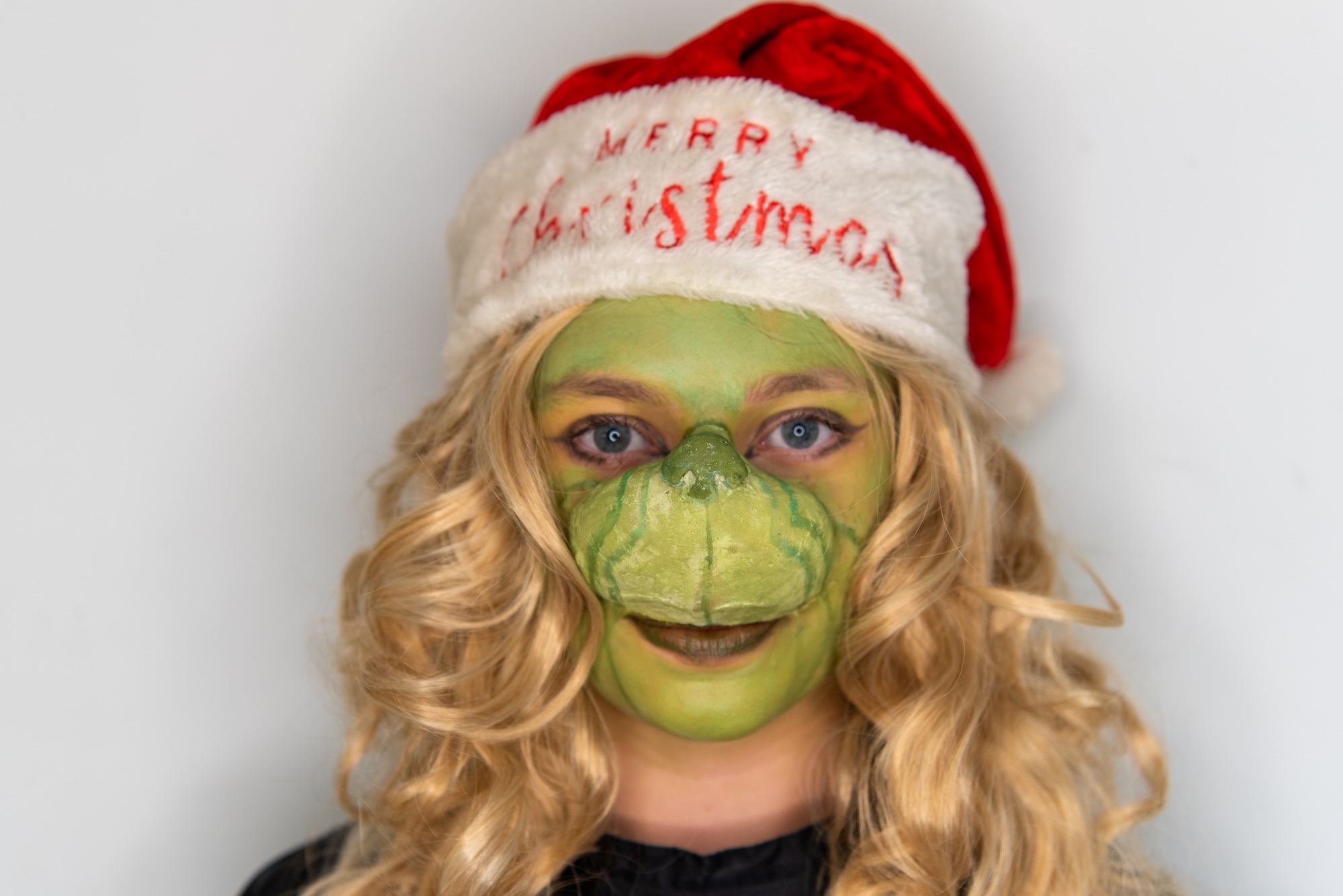 Person with blonde curly hair wearing red and white Santa hat with 'Merry Christmas' text, green face paint covering entire face