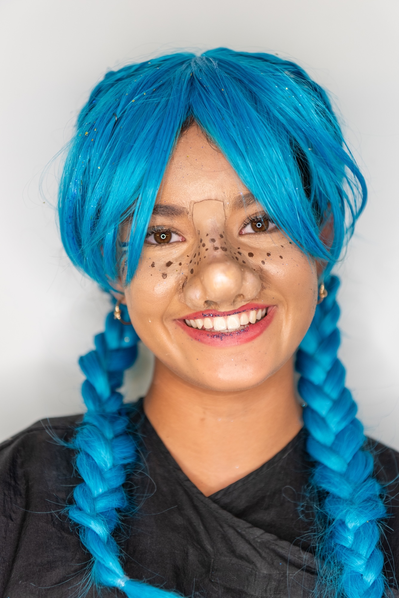 Woman with bright blue braided wig, black dots painted on cheeks and nose, smiling, wearing black top against grey background