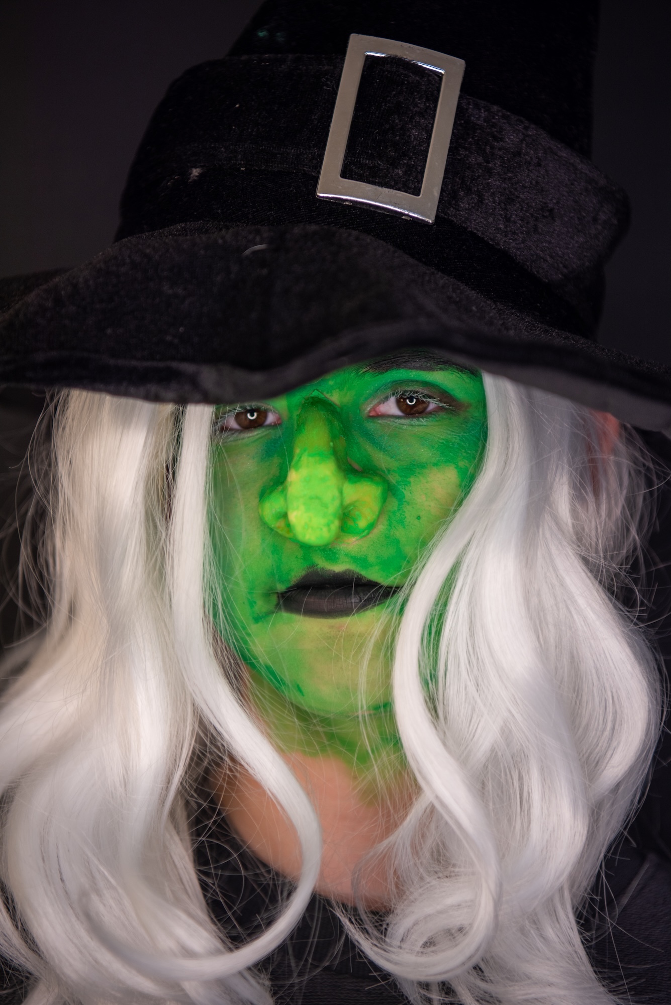 Person wearing black hat with silver buckle, green face paint, and long white wig