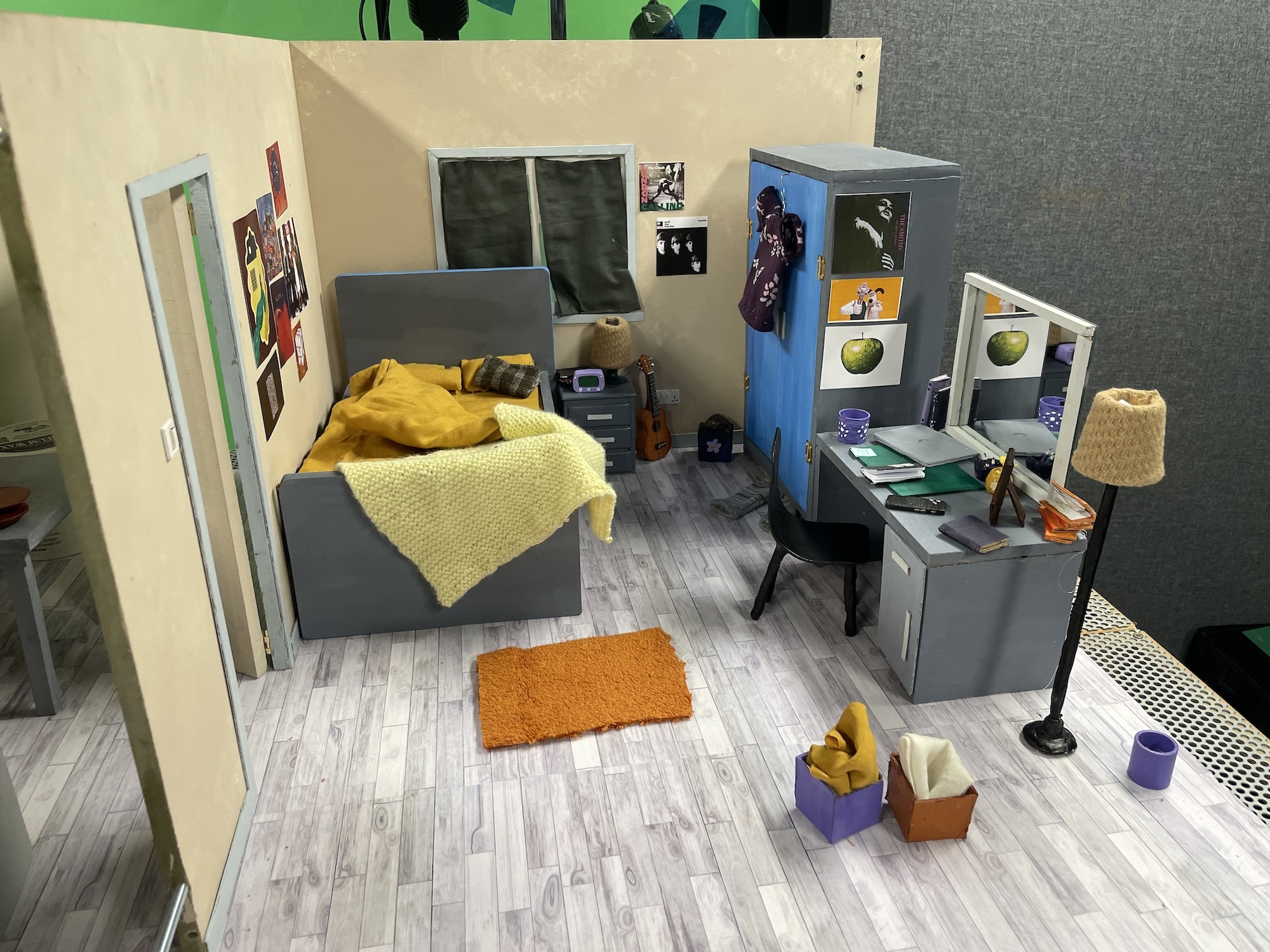 Miniature set for a stop motion animated film of a bedroom, with a messy bed with posters and a window above it, a wardrobe, desk, mirror and lamp on the other side.