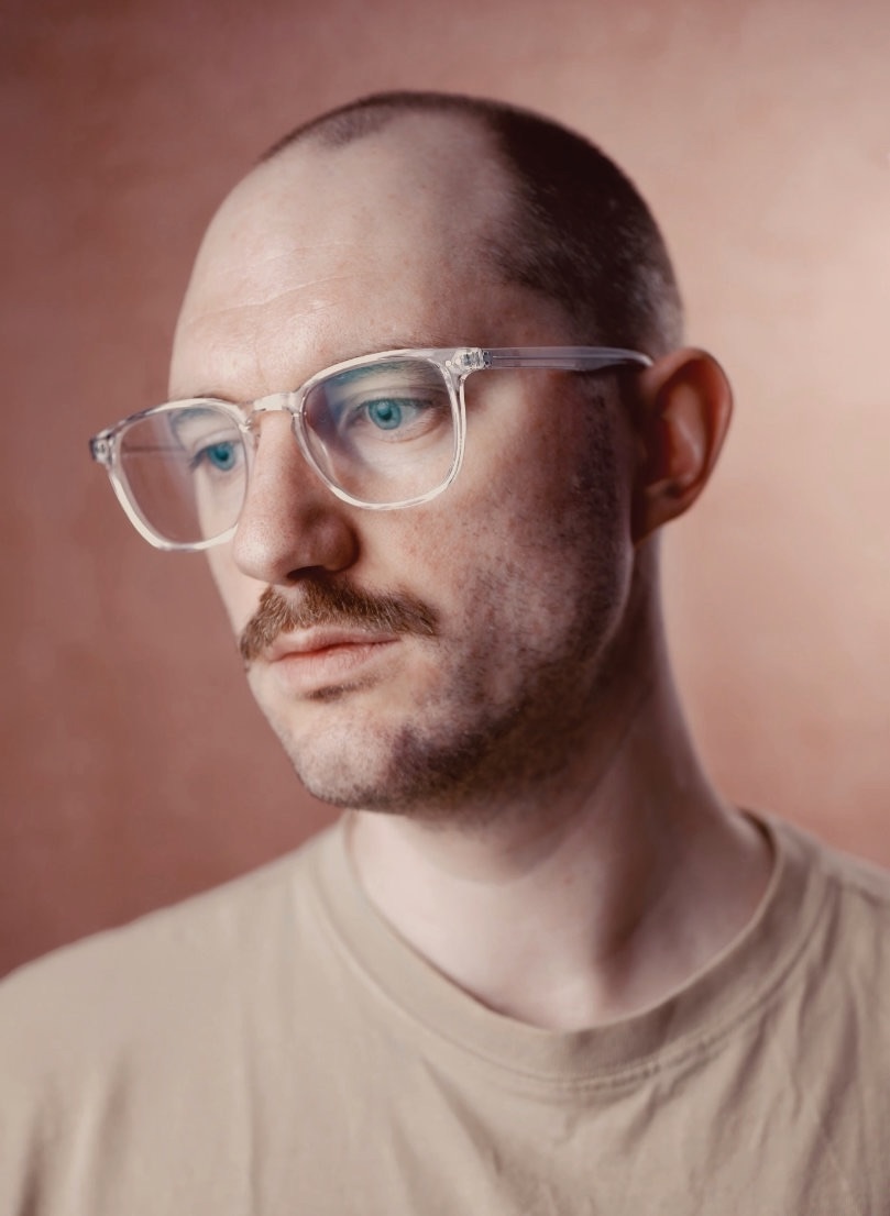 A man with a shaved head wears transparent glasses and a beige t-shirt. He has a light moustache and is facing slightly to the side, with a blurred background of a warm-toned wall.