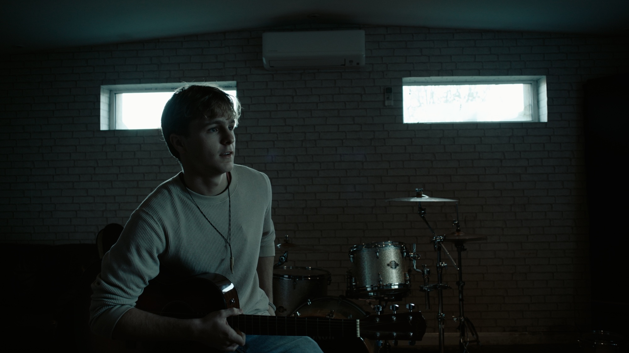 Woman in white jumper sits at dimly lit desk with laptop, two bright rectangular windows behind her casting light into dark room.