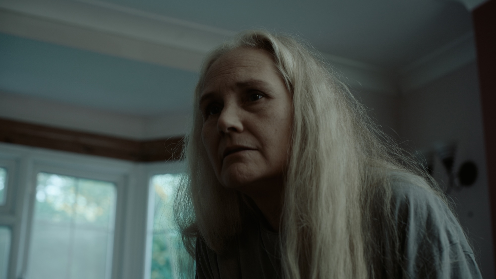 Woman with long blonde hair and pale complexion in dimly lit room with windows showing greenery outside.