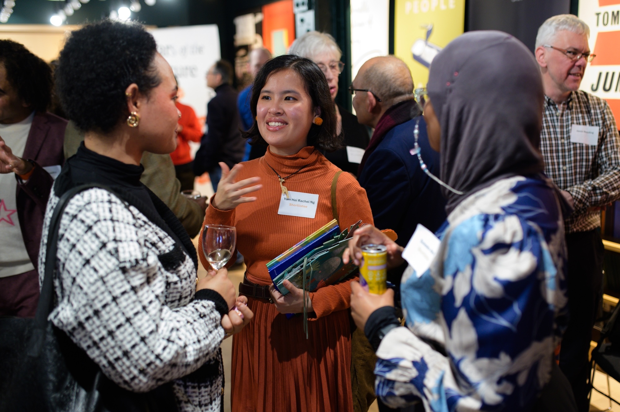 R Ng with two other people in conversation at networking event, one wearing orange dress with name tag, another in chequered jacket holding wine glass.