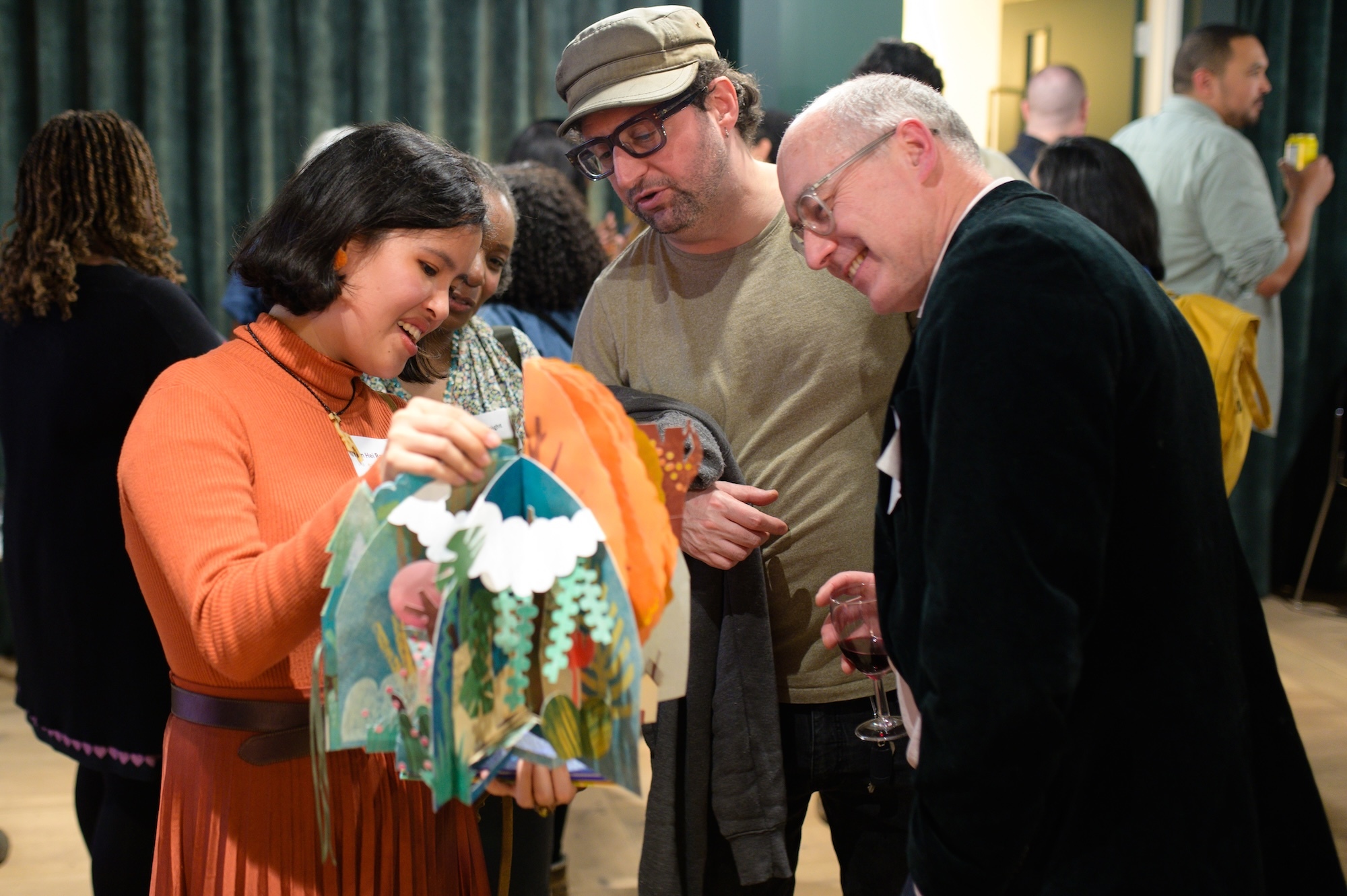 R Ng holding colourful children's book whilst two people in hat and suit look on in indoor setting.