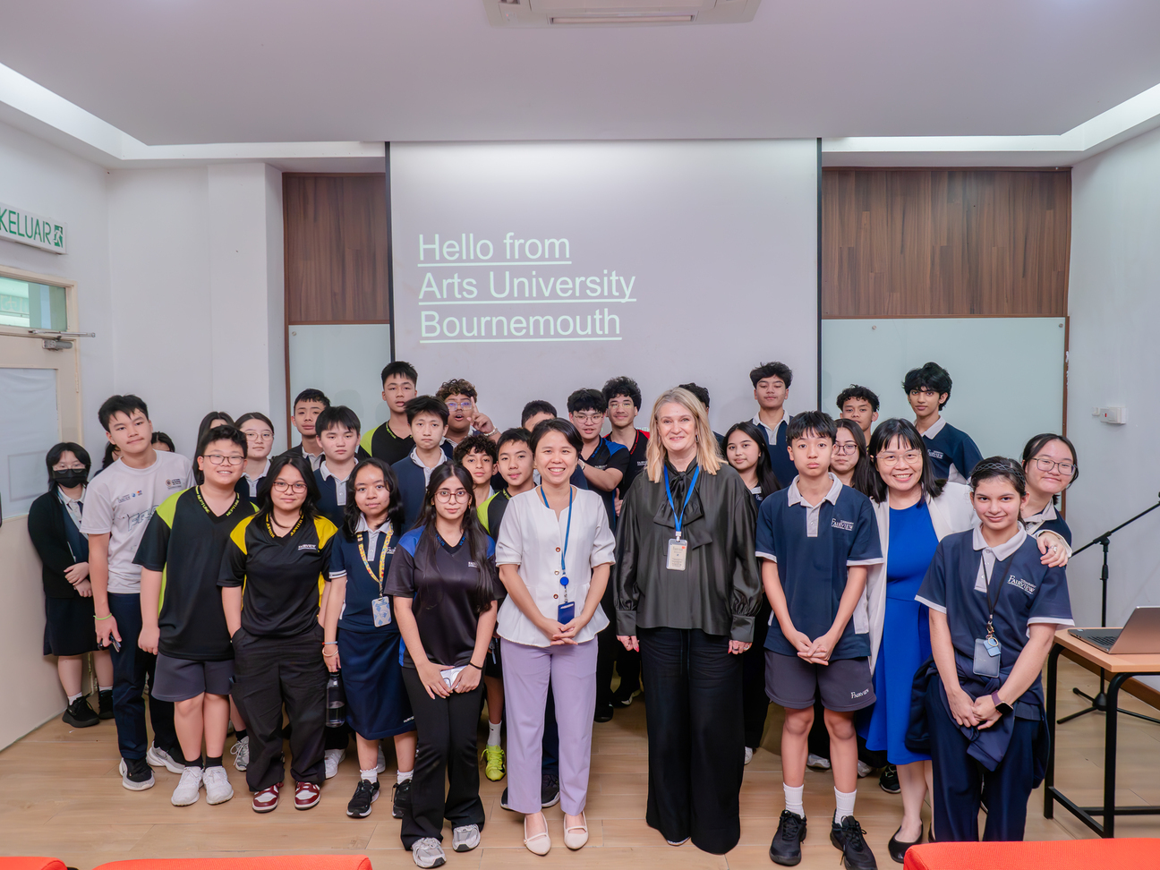 Group of students and staff posing in lecture hall with "Hello from Arts University Bournemouth" projected on screen behind them.