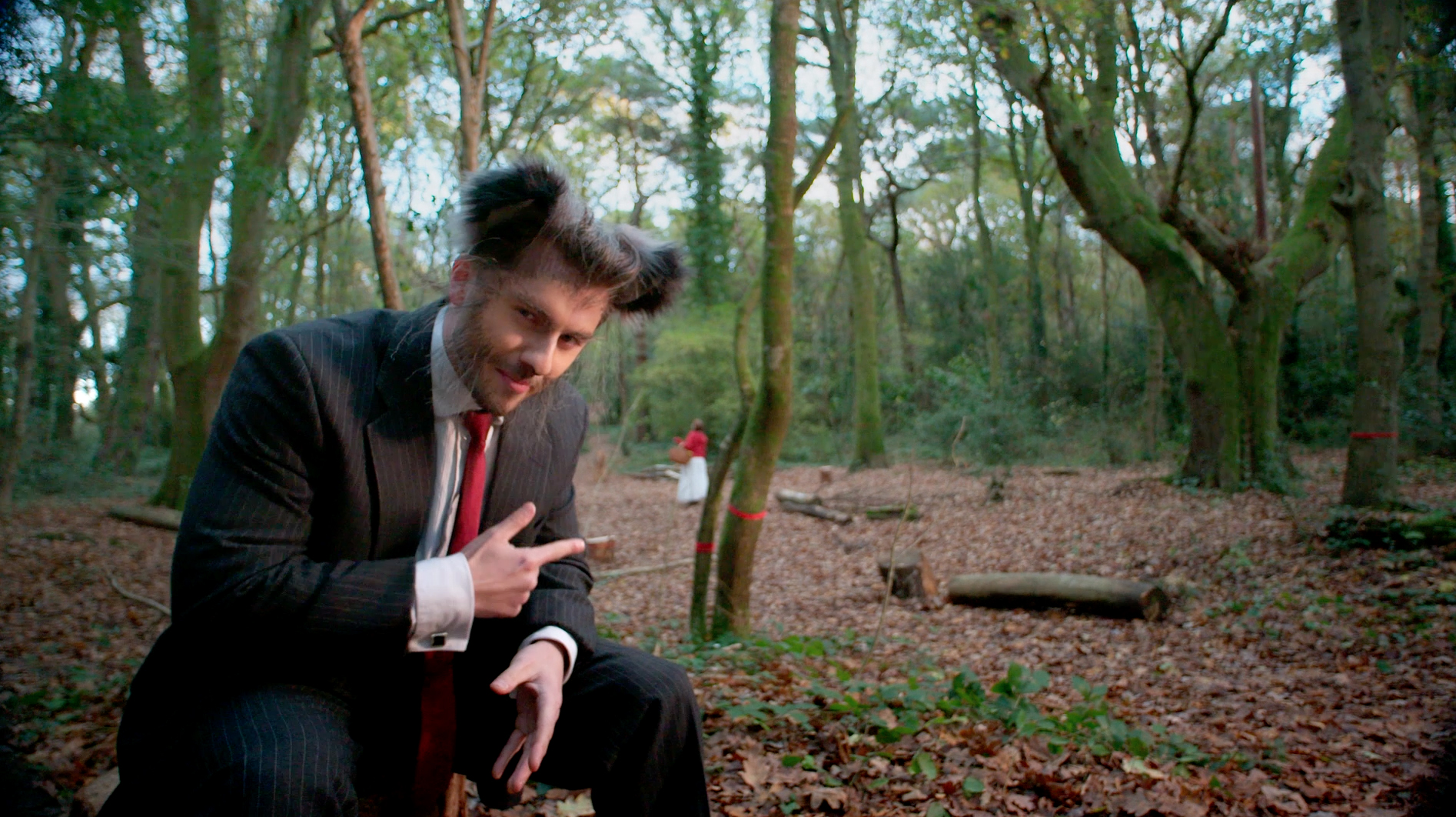 Man in suit with wolf-like prosthetics in a green forest, smirking and pointing at little red riding hood in the distance.