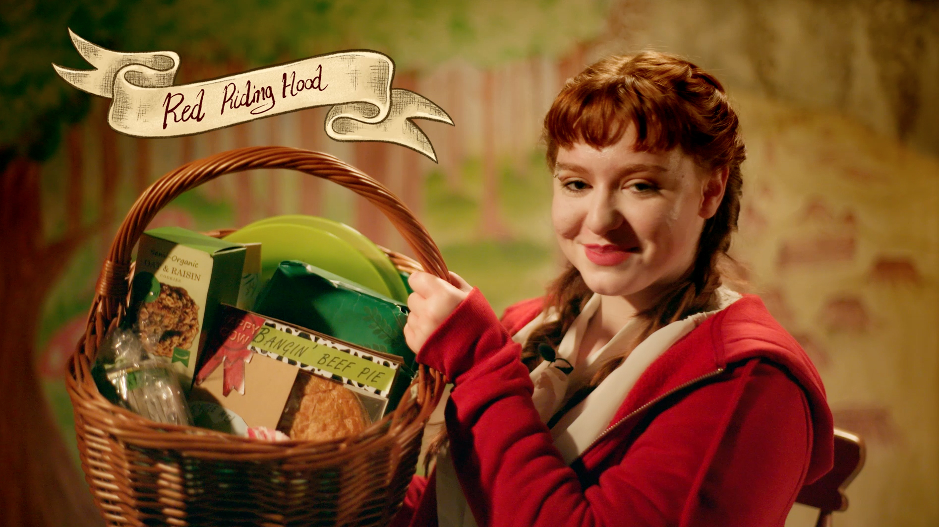 Girl in red hoodie holding a hamper with food items in packaging, text reading 'Red Riding Hood' above.