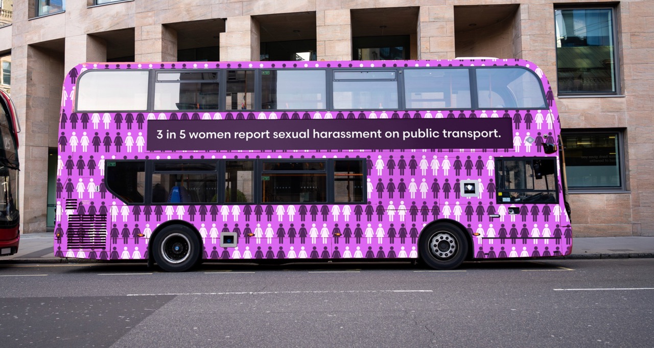 A pink and blue double-decker bus parked on a street. The side of the bus displays a message stating "3 in 5 women report sexual harassment on public transport" along with a pattern of stylised figures.