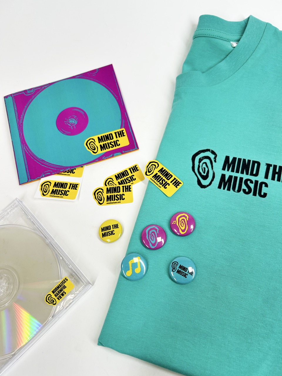 A turquoise t-shirt with the text "MIND THE MUSIC" and a colourful CD with a spiral design are displayed alongside yellow stickers, each with the same text, and several buttons featuring similar designs on a white surface.