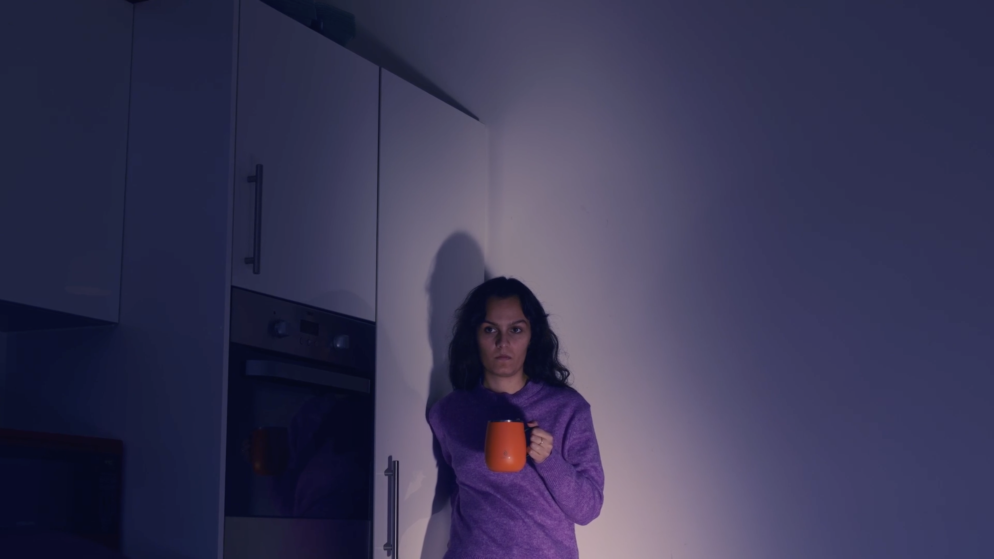Film still of a woman looking serious, holding a large orange coffee mug in one hand, kitchen cabinets and a large white wall behind her in the dark background.