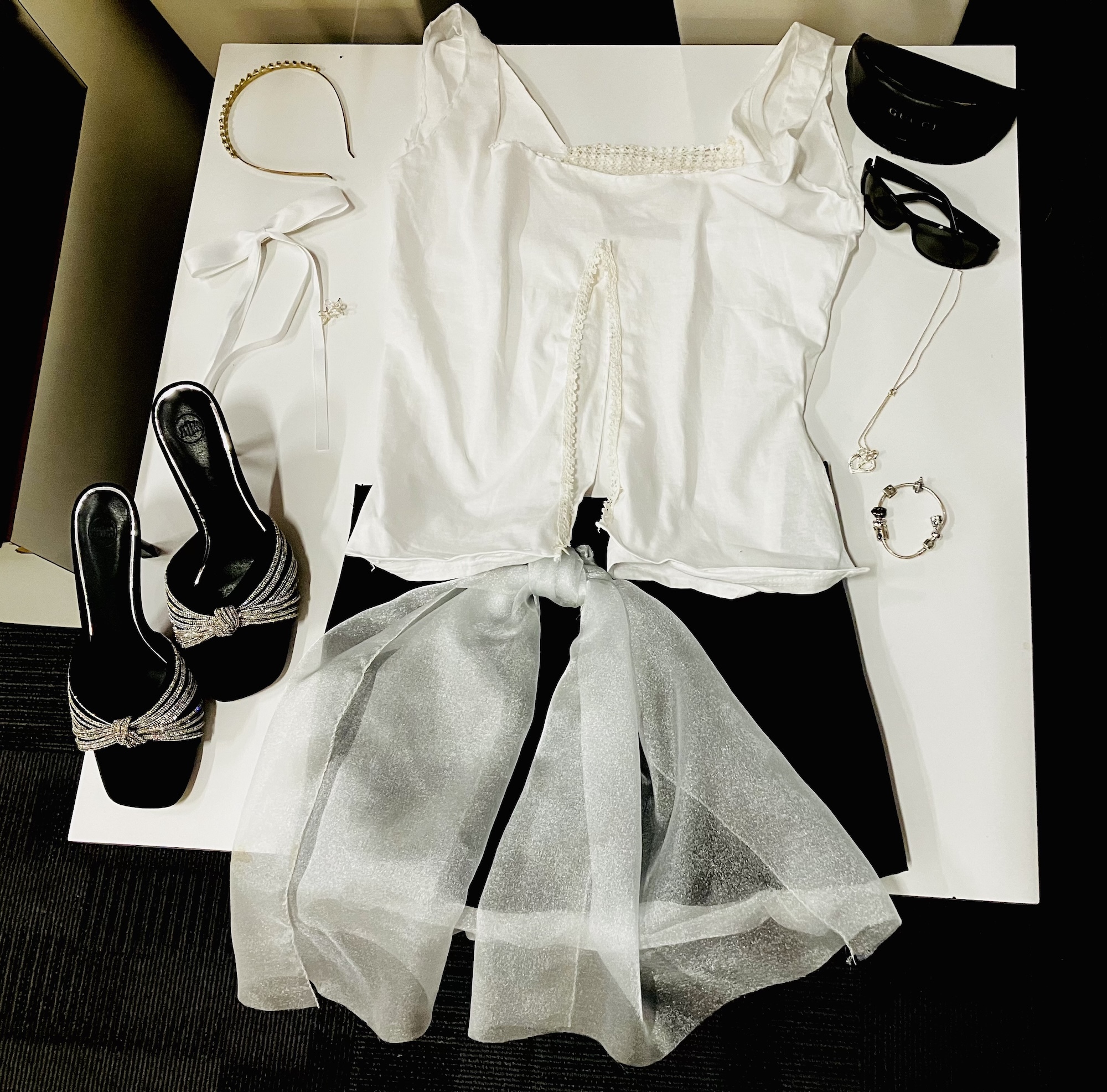 White outfit laid flat: sleeveless top, sheer skirt, black strappy heels, sunglasses, and silver jewellery arranged on beige surface