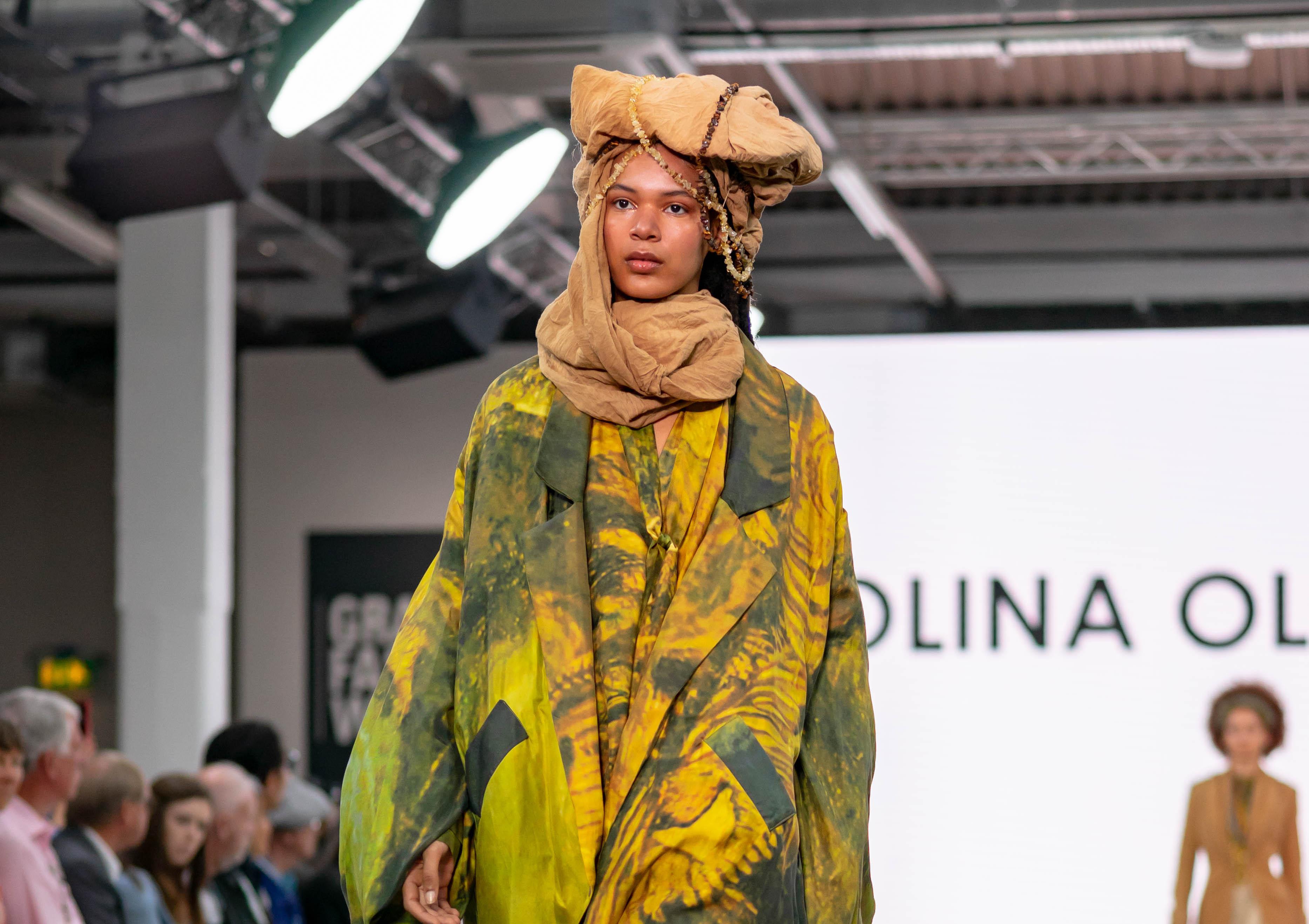 Model in a colourful outfit and headdress walks down a runway as part of a fashion show.