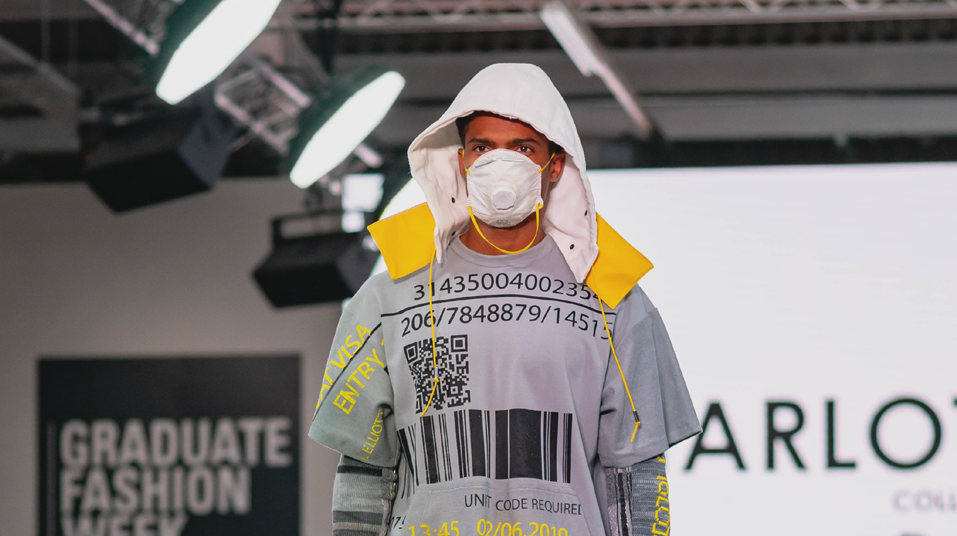 A model wearing a hood, a face mask and a T-shirt with various bar codes and a QR code printed on it, walks down a runway