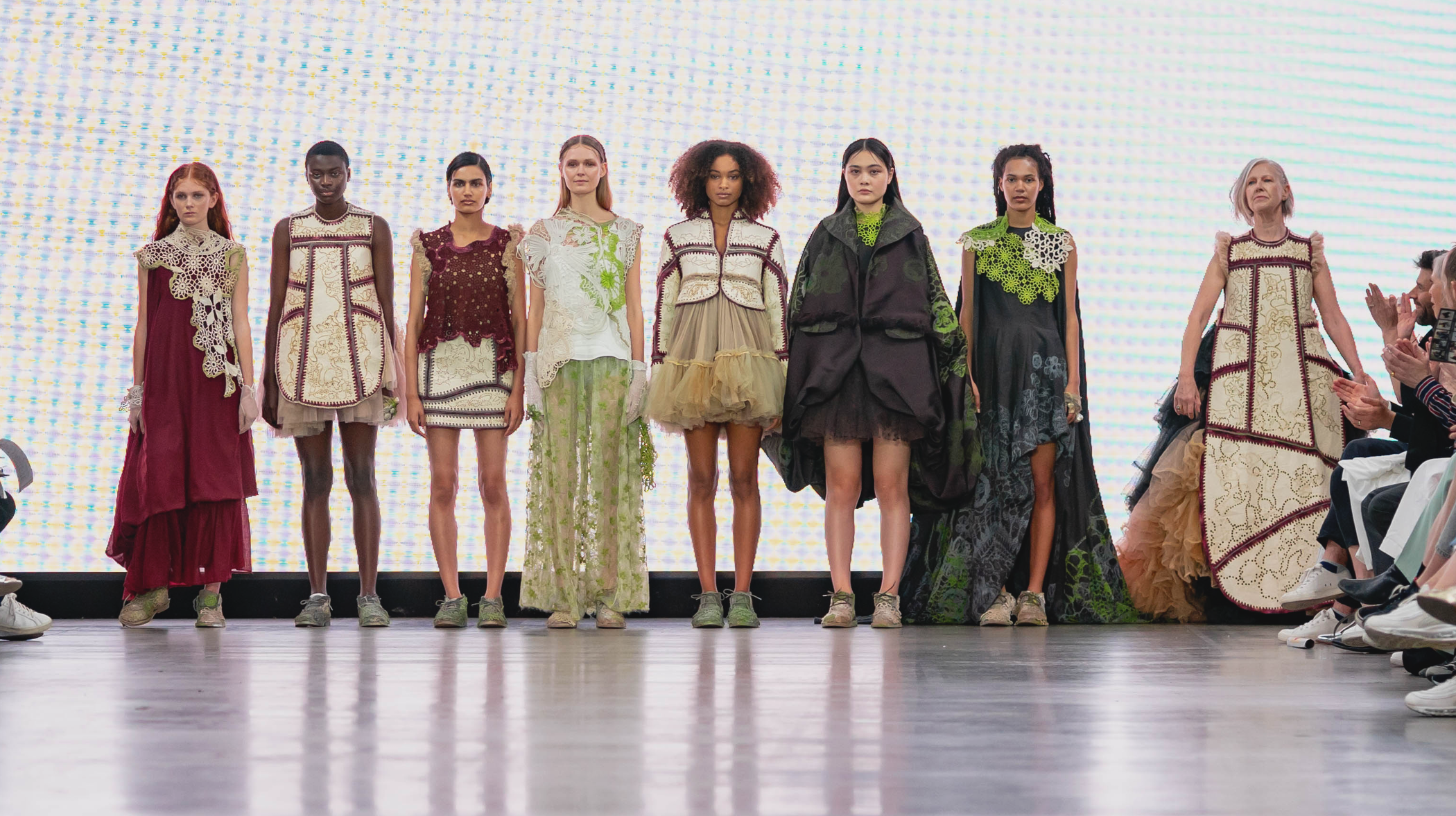 Eight people line up in different outfits against a bright backlit screen in the middle of a fashion show.