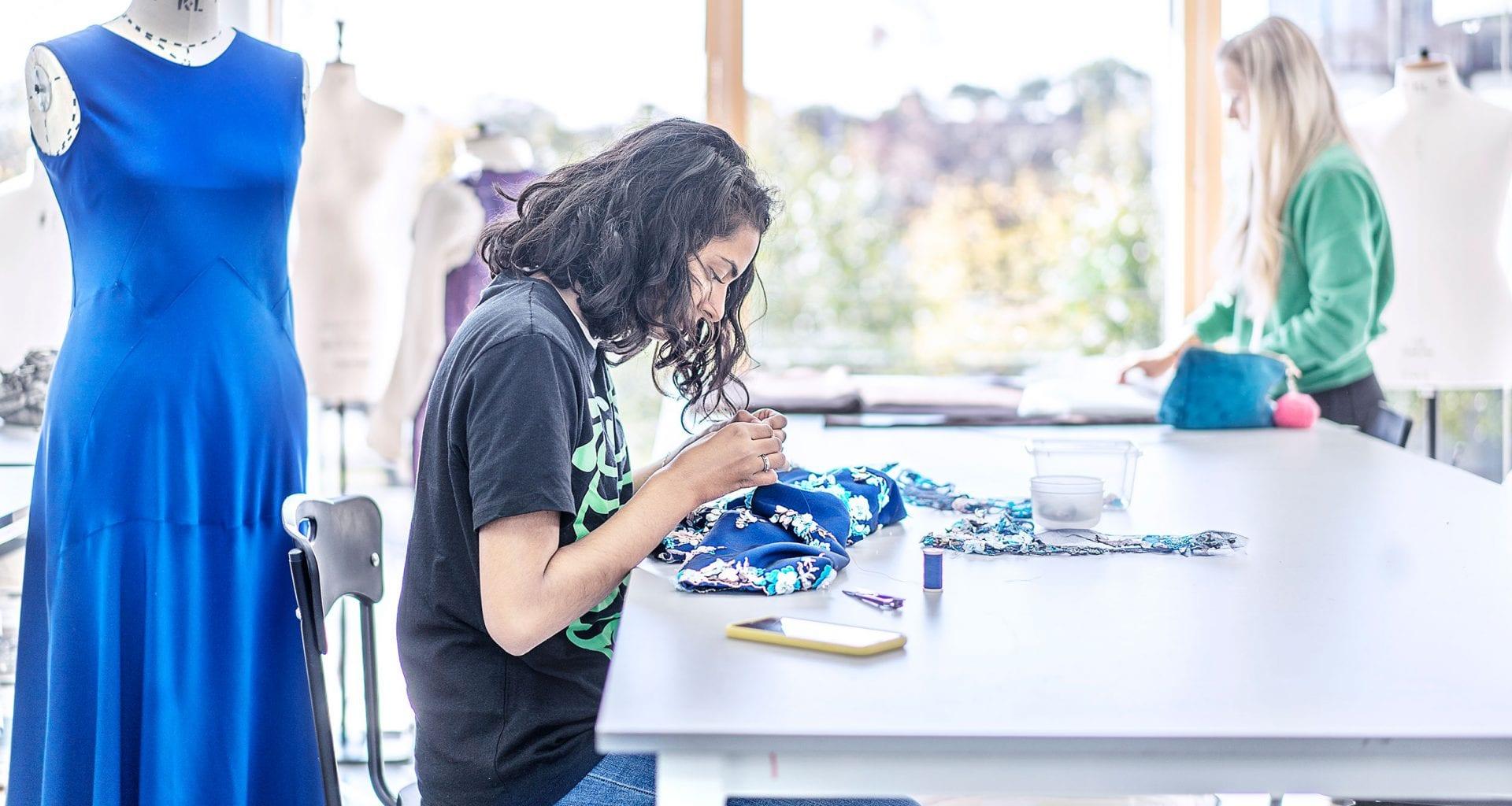 Woman wearing a black t-shirt sat at a desk sewing blue pattered material. There's a blue dress on a mannequin behind her