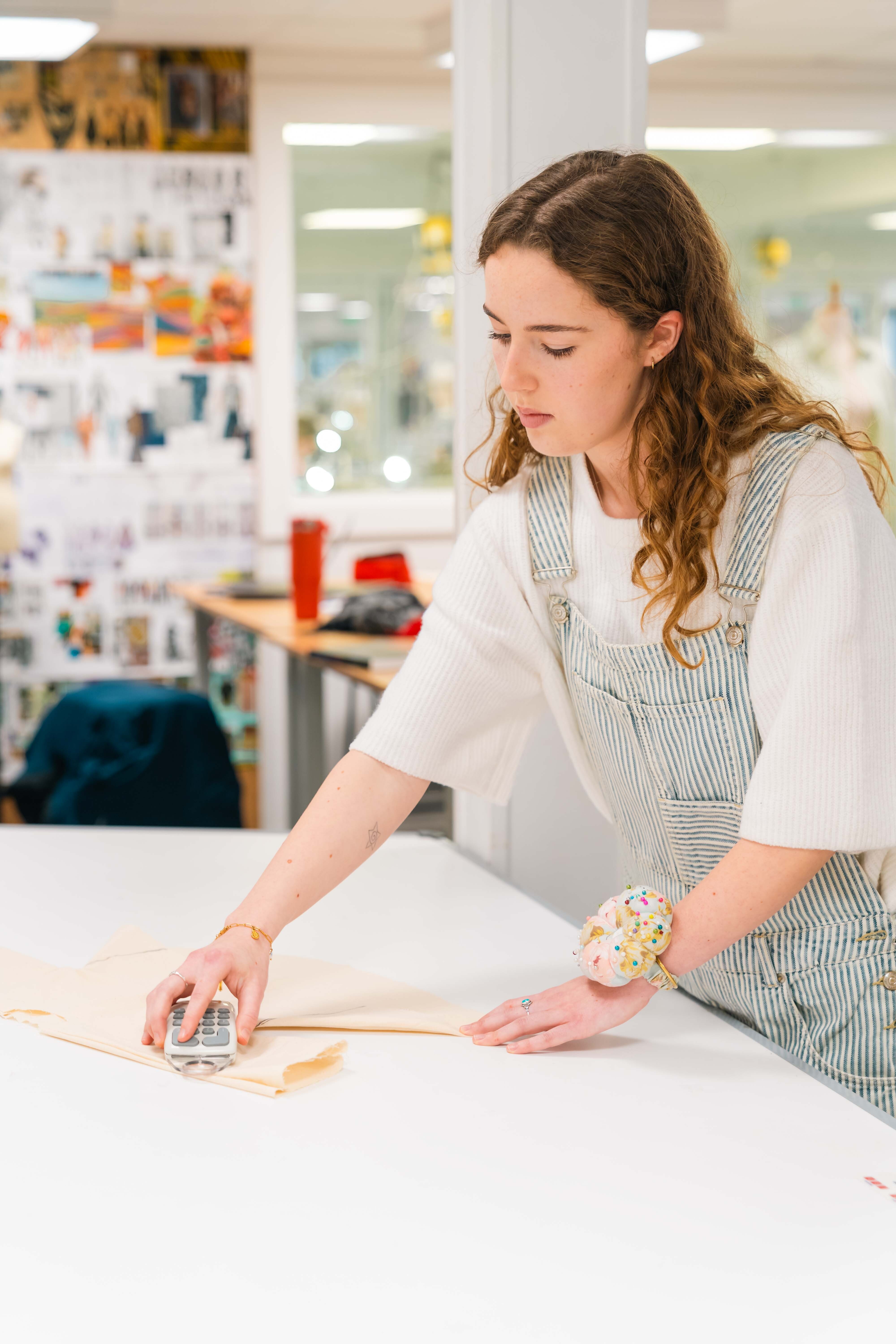 A BA (Hons) Fashion student in striped apron and white top using roller on paper at white table in bright workspace with colourful artwork on walls.