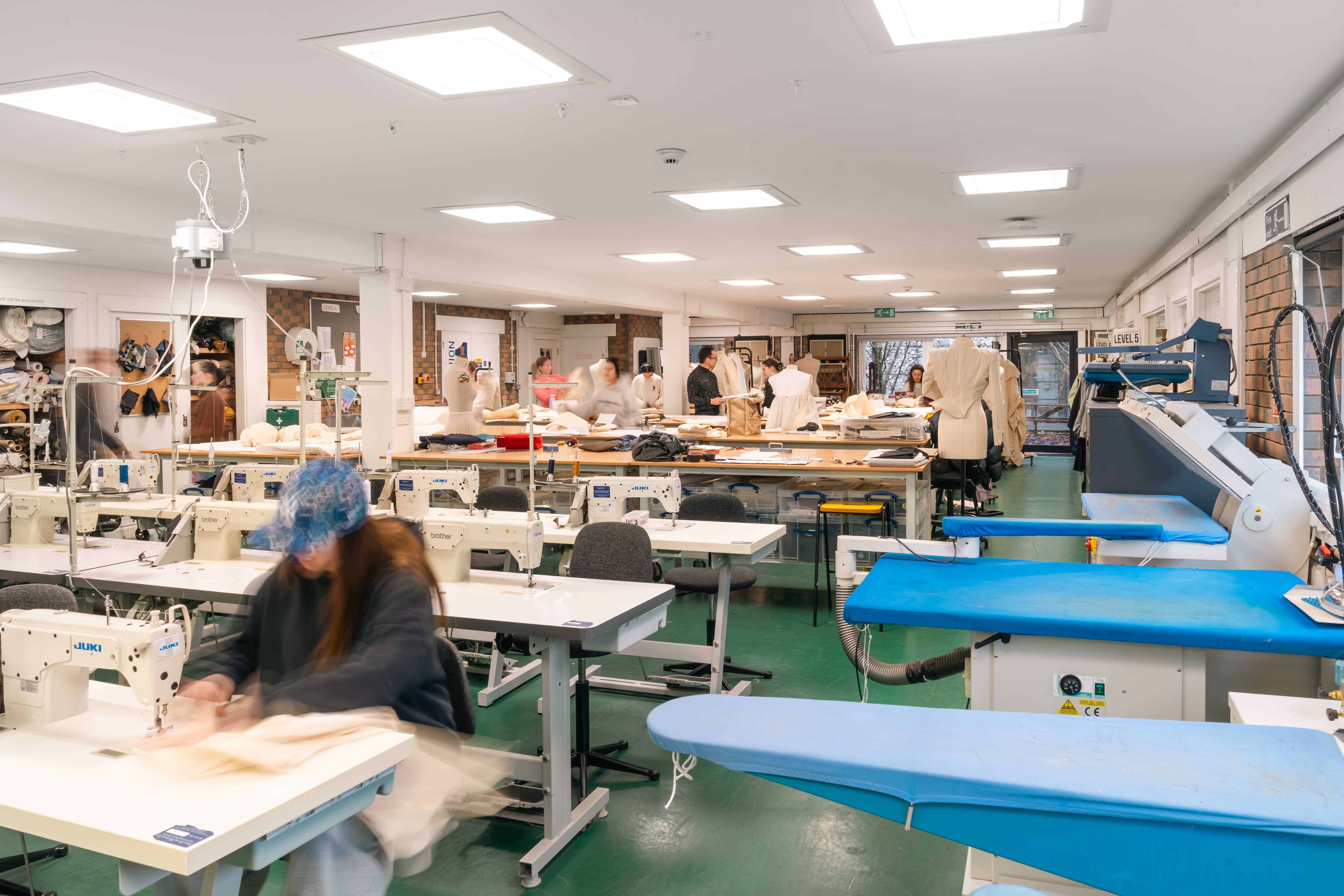 Bright art classroom with white tables, blue chairs, and green flooring. Students work at stations with artwork displayed on walls.