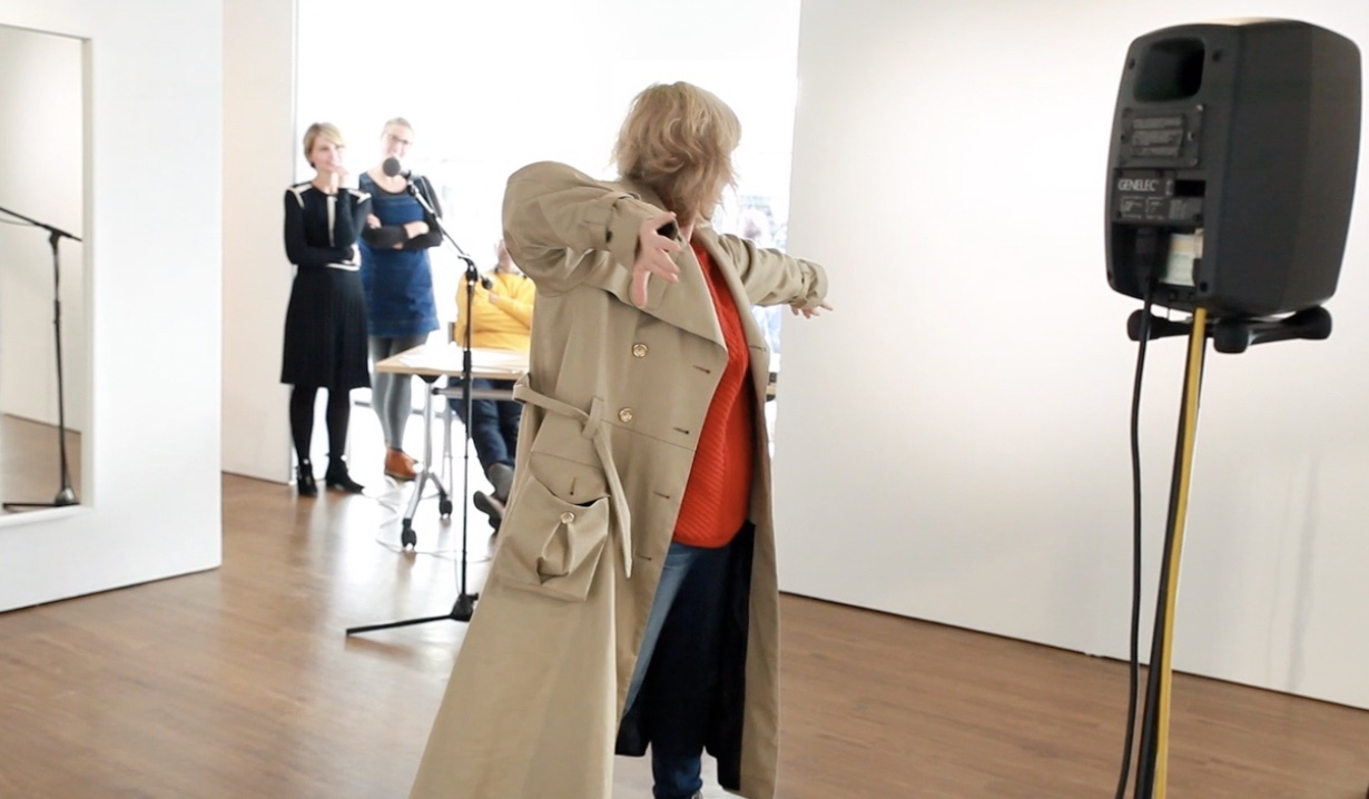 A person wearing a trench coat poses with their back to the camera towards a white wall, next to a speaker. Two other people watch from the background.