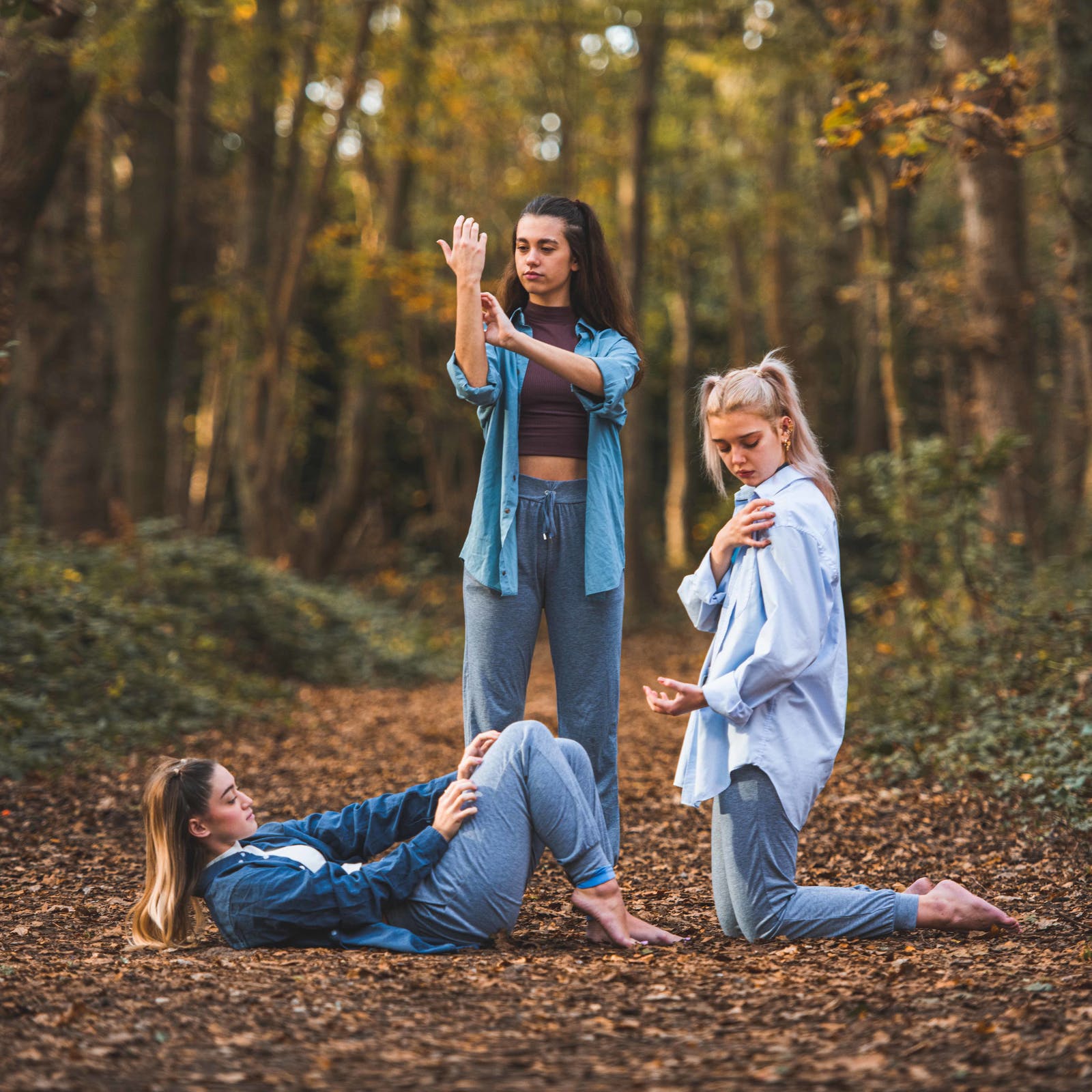 Three women practise yoga poses on a forest path, with autumn trees and fallen leaves in the background.