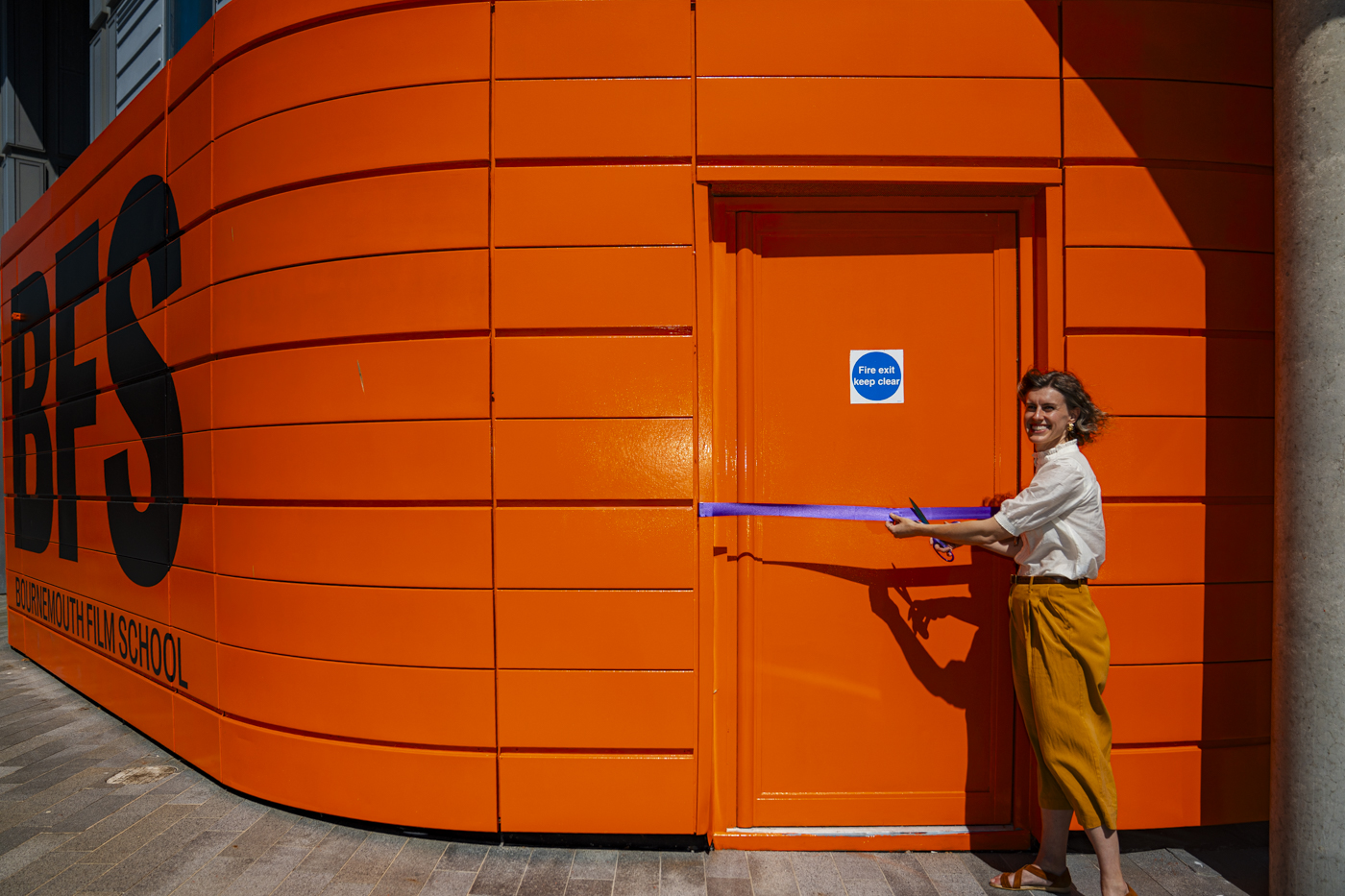 A woman with curly hair in a cream blouse and mustard-yellow trousers stands beside an orange door with a blue sign that reads 'Fire Exit.' She is cutting a blue ribbon in front of a building featuring large black lettering stating 'BFS'.