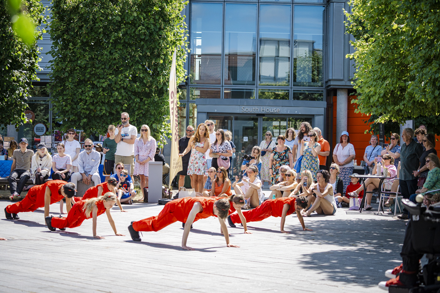 A group of performers in red costumes are engaged in a dance routine on a paved area surrounded by an audience sitting and standing under trees. The background features a modern building with large windows.