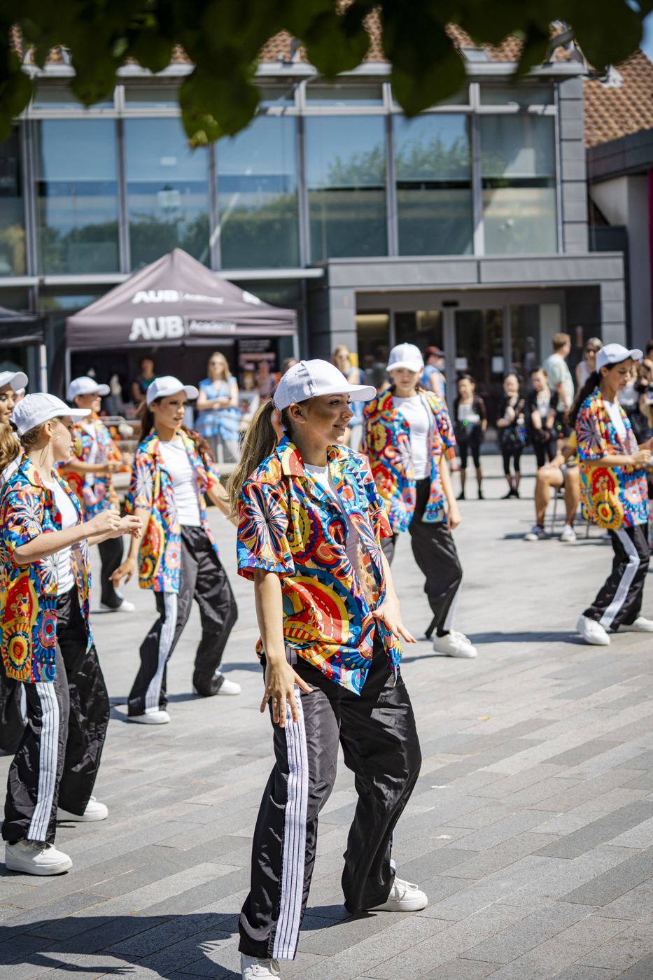 A group of dancers performs in a plaza, wearing vibrant patterned shirts and black athletic pants. The dancers are positioned in a line, with a backdrop of modern buildings and people observing in the background.