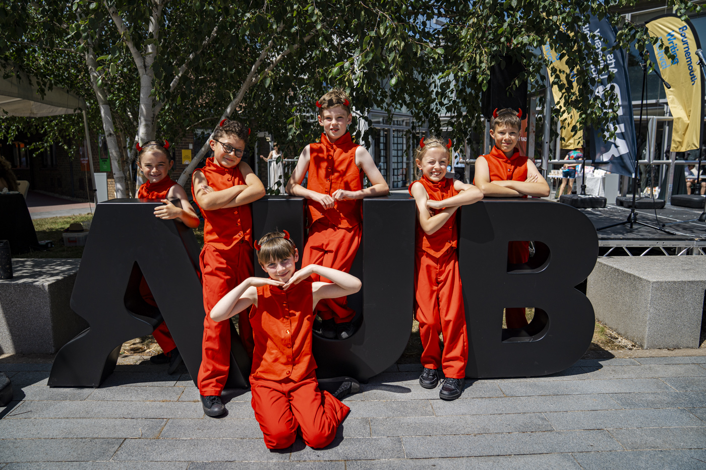Six children wearing bright red outfits pose together around a large black letter formation spelling 'AUB'. The setting is outdoors with trees and a building in the background.