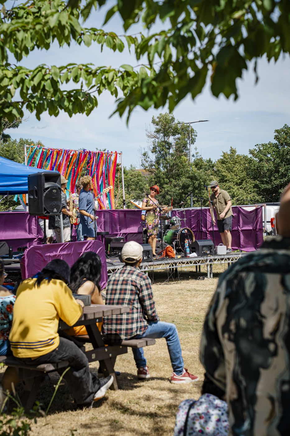 A small outdoor stage features a band performing with microphones and instruments. In the foreground, people are seated at picnic tables, while colourful ribbons drape behind the band, and trees provide shade in a park setting.
