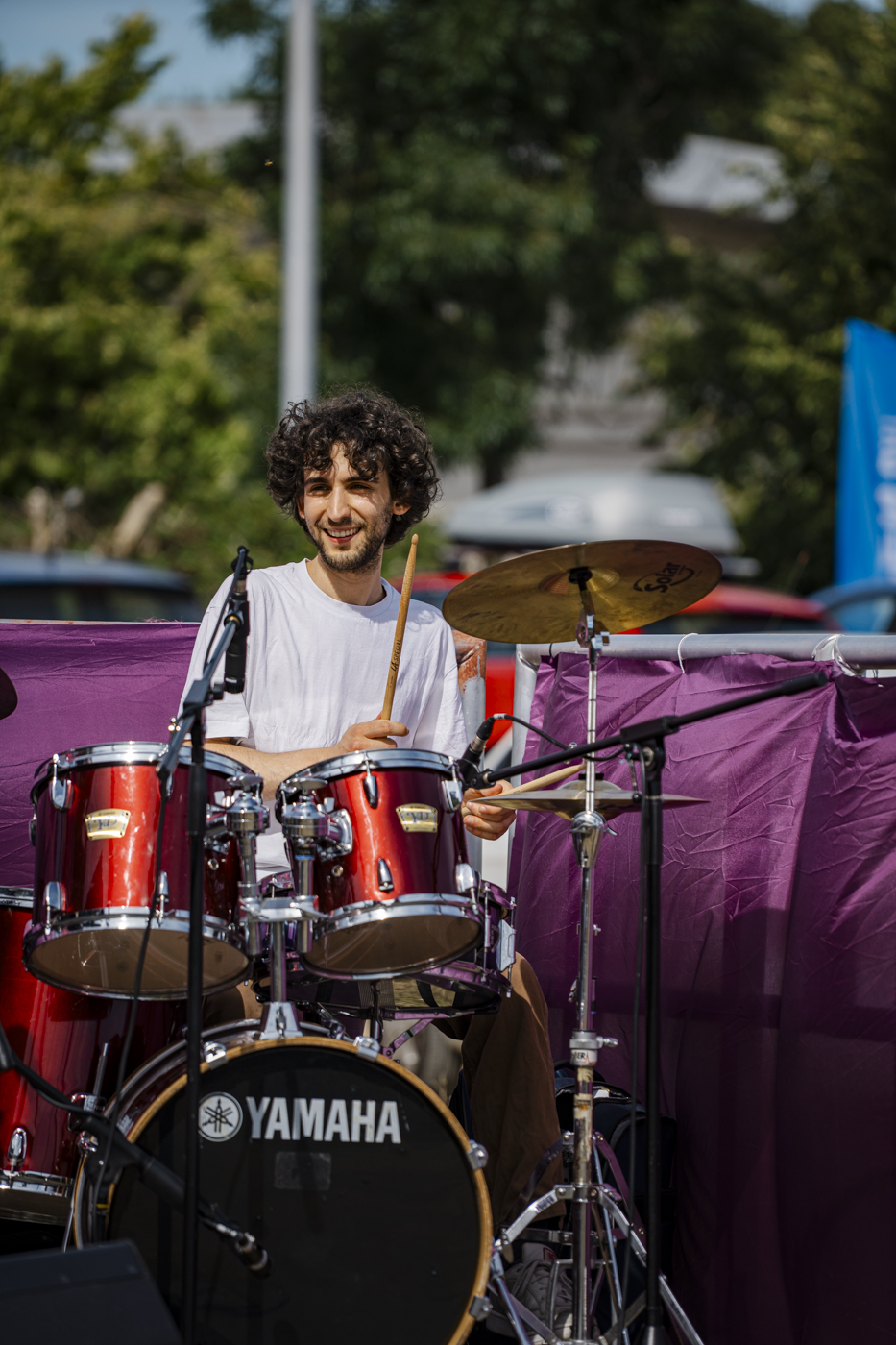 A young man with curly hair plays a red drum kit outdoors during a performance. He is wearing a white t-shirt, holding drumsticks, and sitting behind the drums, with a backdrop of trees and parked cars.