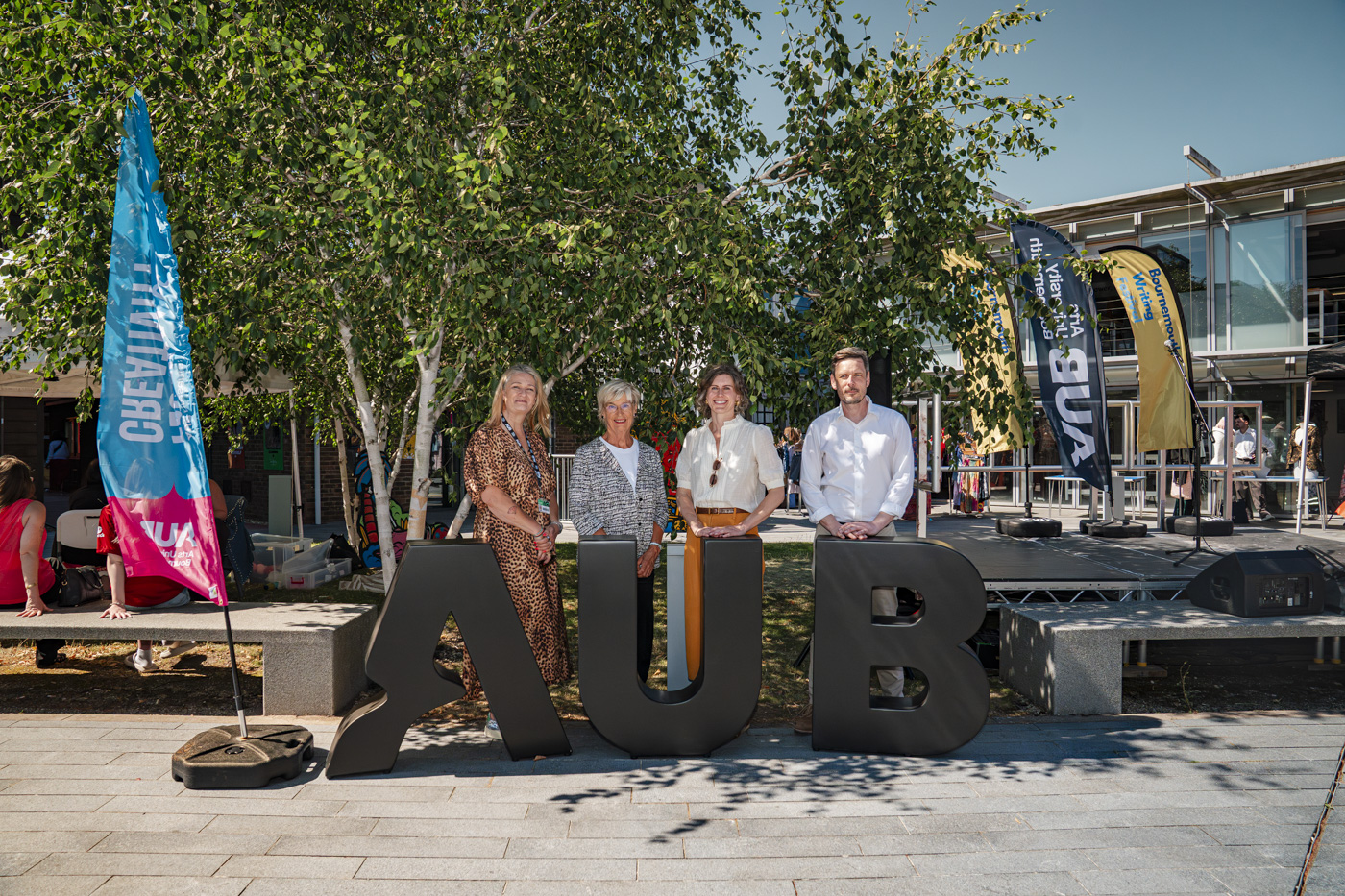 Four individuals stand next to a large black letter 'AUB' in an outdoor setting, surrounded by greenery and modern buildings. Behind them, flags and signs add color to the scene, indicating an event.