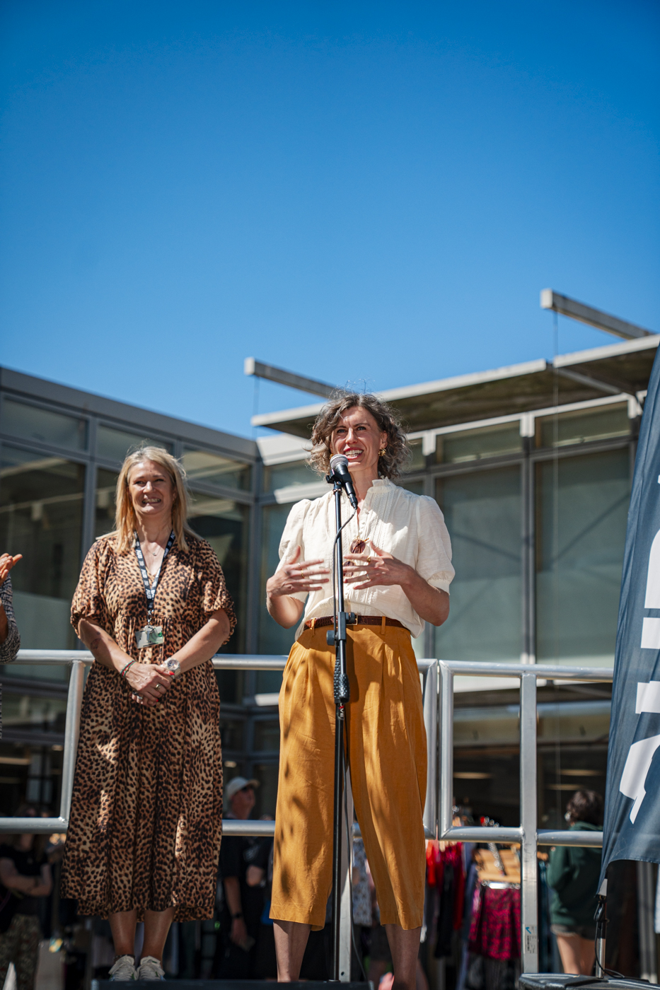 A woman in a white blouse and yellow trousers stands at a microphone, addressing an audience outdoors. To her left, another woman in a brown patterned dress is smiling, while a crowd is visible in the background under a clear blue sky.