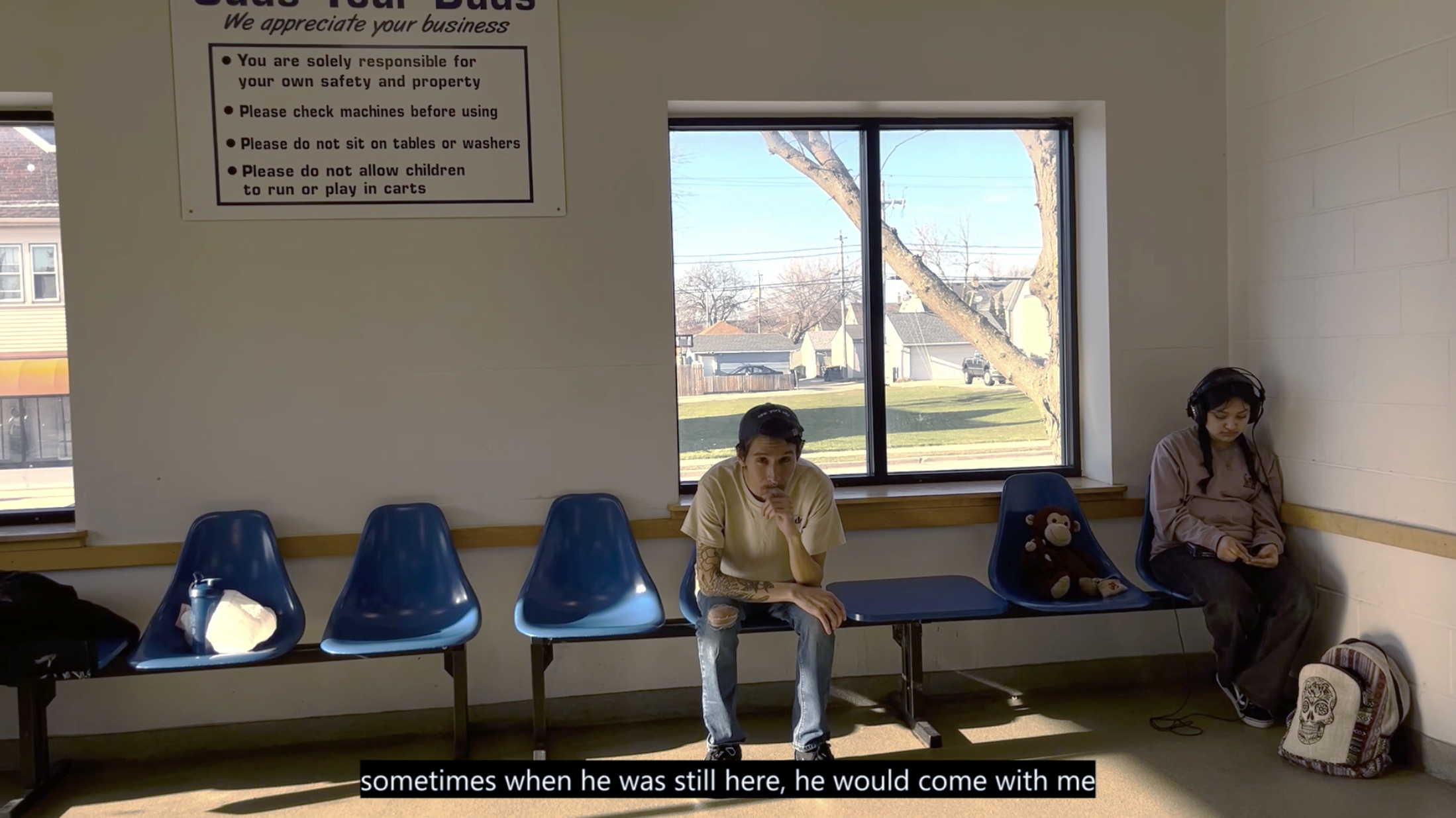 Waiting room with blue plastic chairs, two people seated apart, white walls, window showing construction crane outside, safety poster visible.