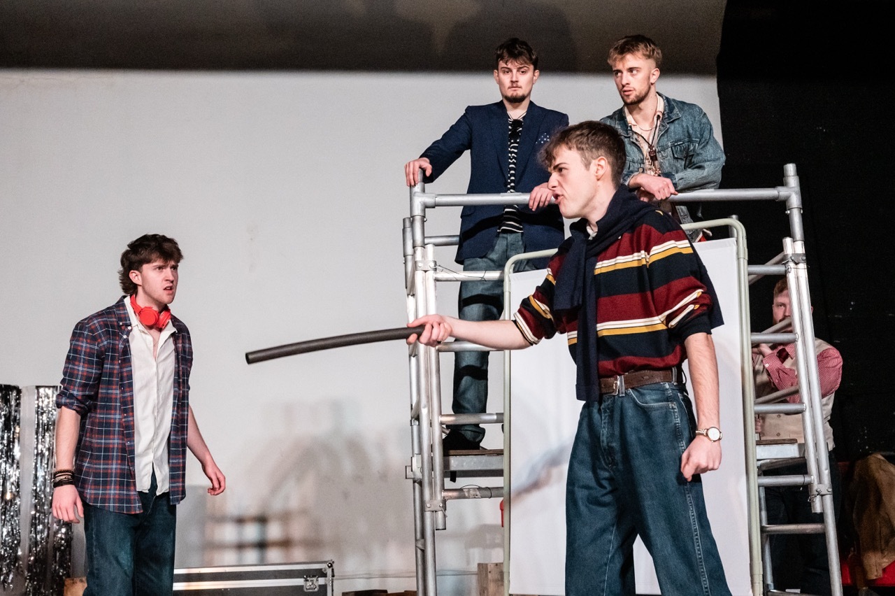 Four male performers on stage with metal scaffolding platform. Two men standing on raised structure, one in foreground wearing striped shirt.